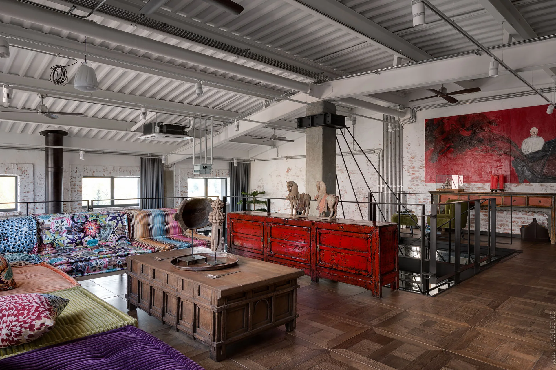 An industrial loft living room with a colorful modular floor sofa, a carved wood trunk coffee table, and a bright red lacquered cabinet.