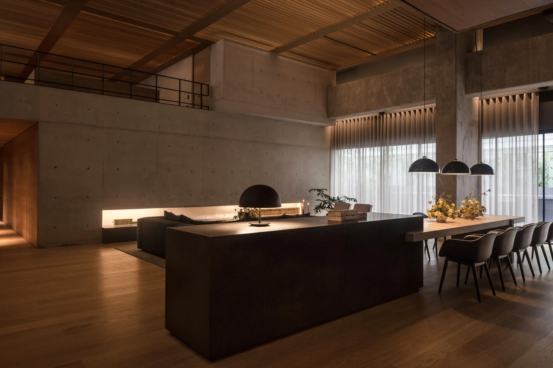 A spacious modern dining area features exposed concrete walls, a dark stone island, a long table with chairs, and a slatted wood ceiling.
