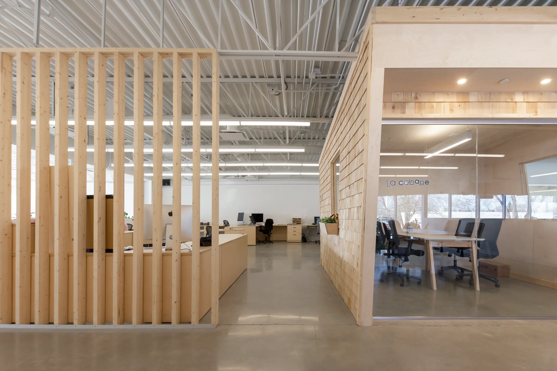 A modern open-plan office space featuring a light wood slat partition, a glass-walled meeting room, and polished concrete flooring under an industrial ceiling.