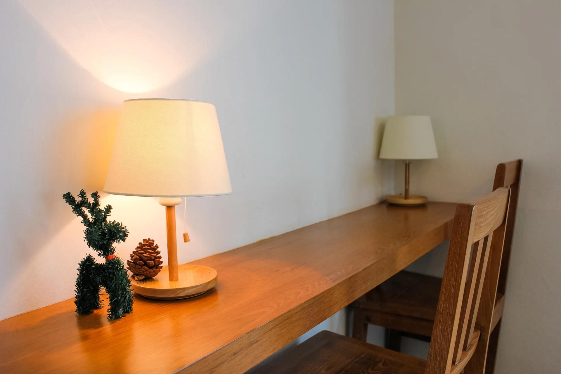 A long wooden desk features two glowing table lamps, a wooden chair, and a small pinecone and green figurine against a plain off-white wall.