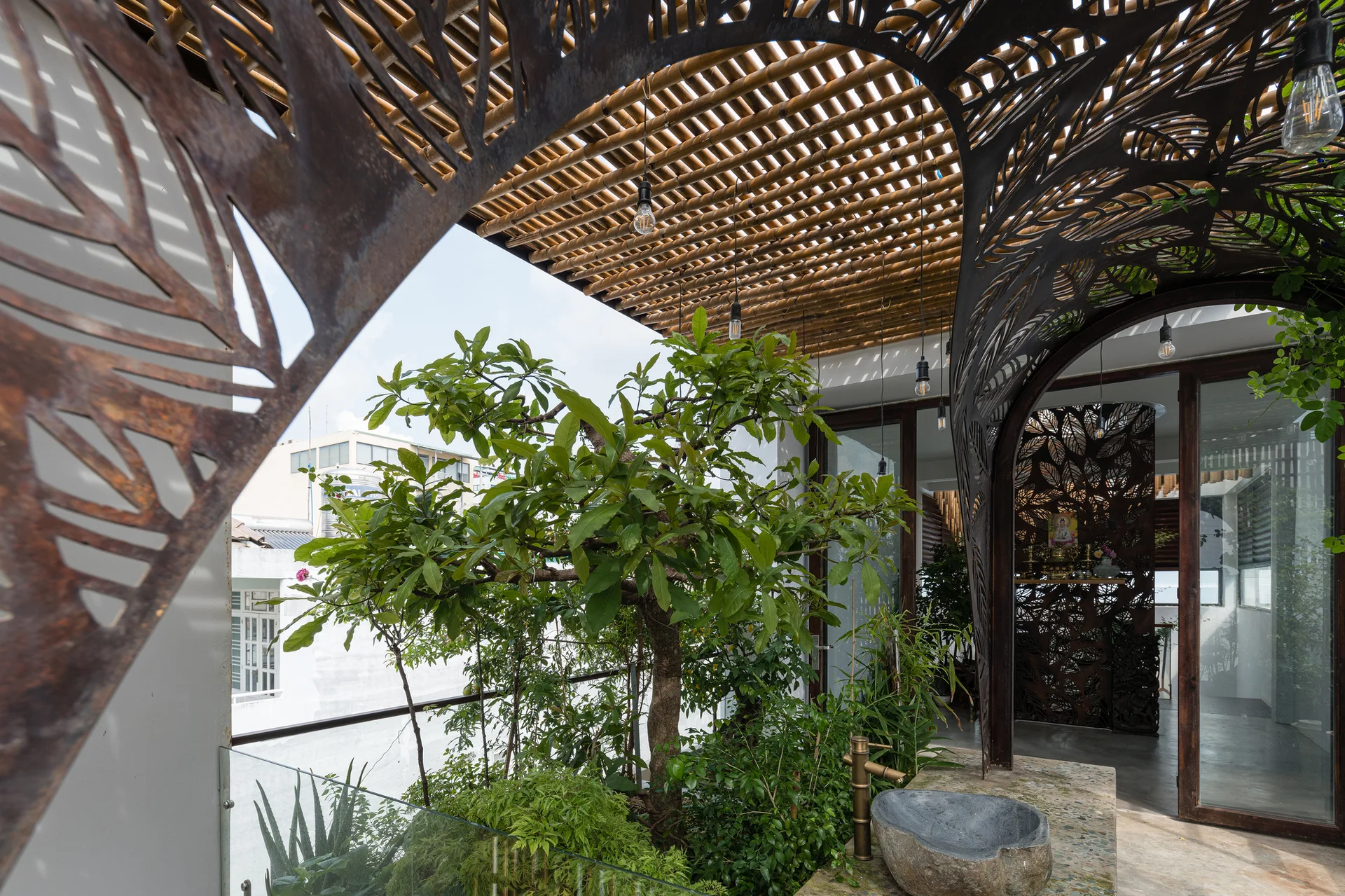 An outdoor rooftop terrace featuring a bamboo slat ceiling, a rusted laser-cut metal archway with leaf patterns, green plants, and a rustic stone sink.