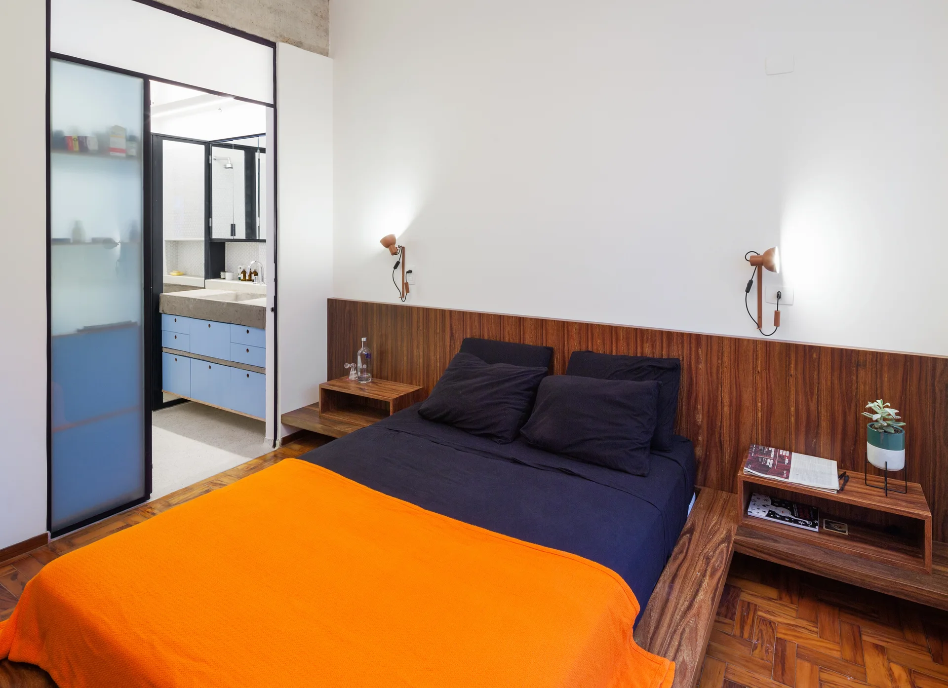 A modern bedroom features an integrated wood headboard with floating shelves, dark bedding, a bright orange blanket, and an adjacent bathroom.