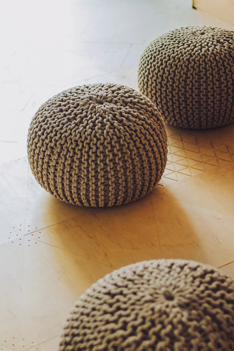 Three round knitted beige poufs rest on a light wood floor featuring geometric triangular and dotted pattern detailing.