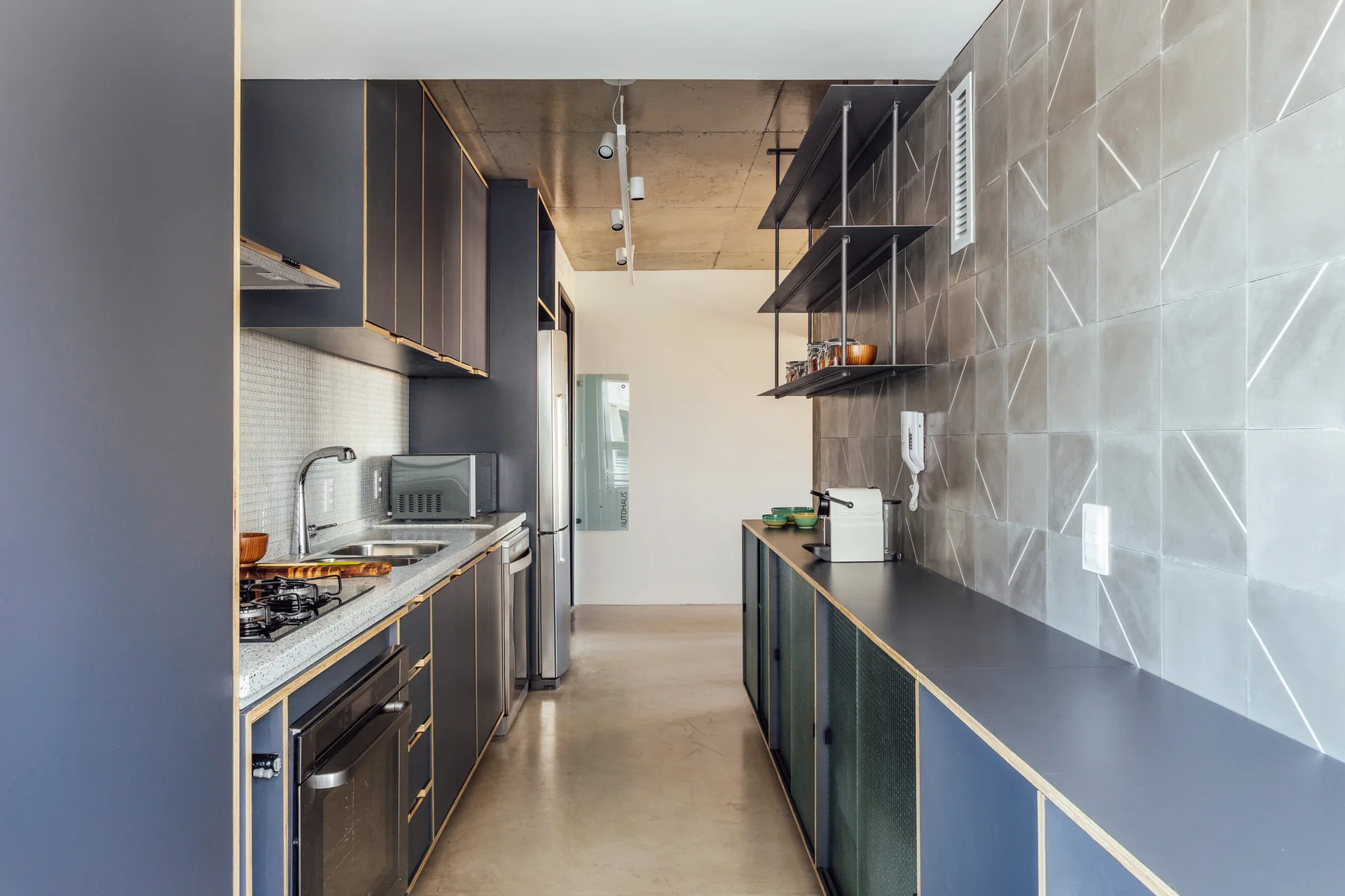 A modern galley kitchen featuring dark grey cabinets with exposed plywood edges, a geometric tiled wall, and a polished concrete floor.