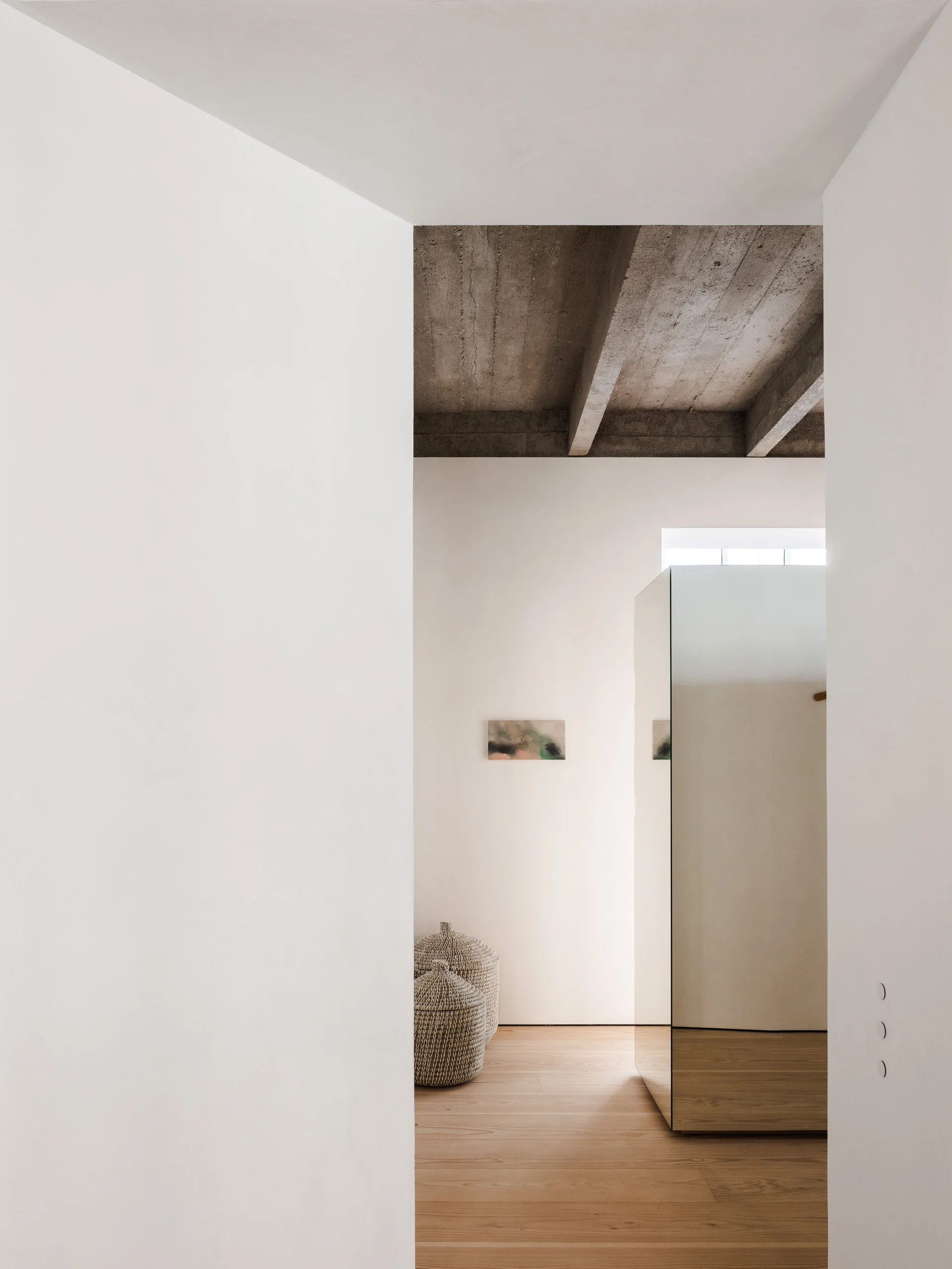 A minimalist hallway viewed through a doorway, featuring a raw concrete ceiling beam, light wood floors, and a tall mirrored column.