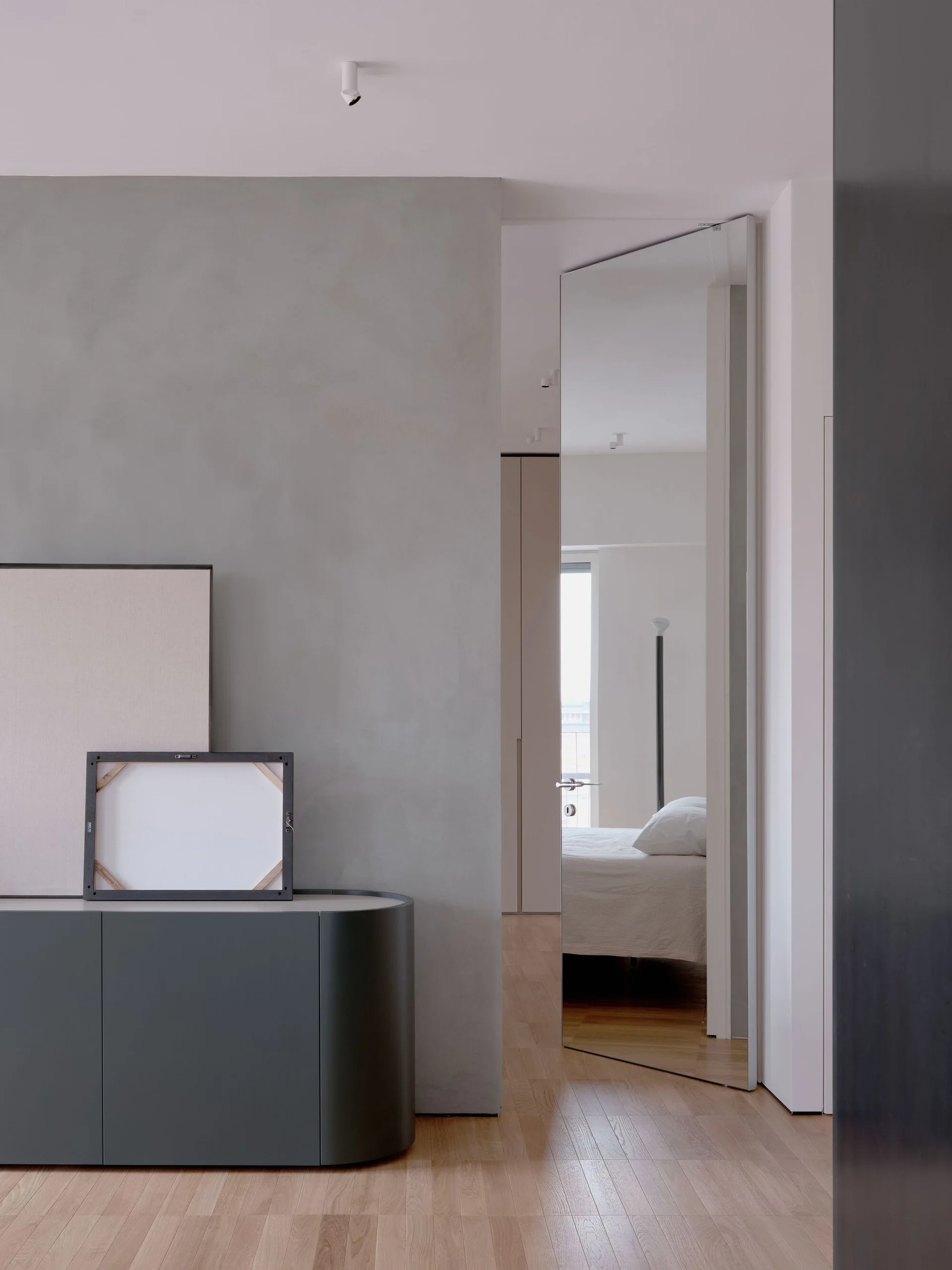 A minimalist hallway featuring a dark gray curved cabinet with leaning frames and a tall mirrored door reflecting an adjacent bedroom with light wood floors.