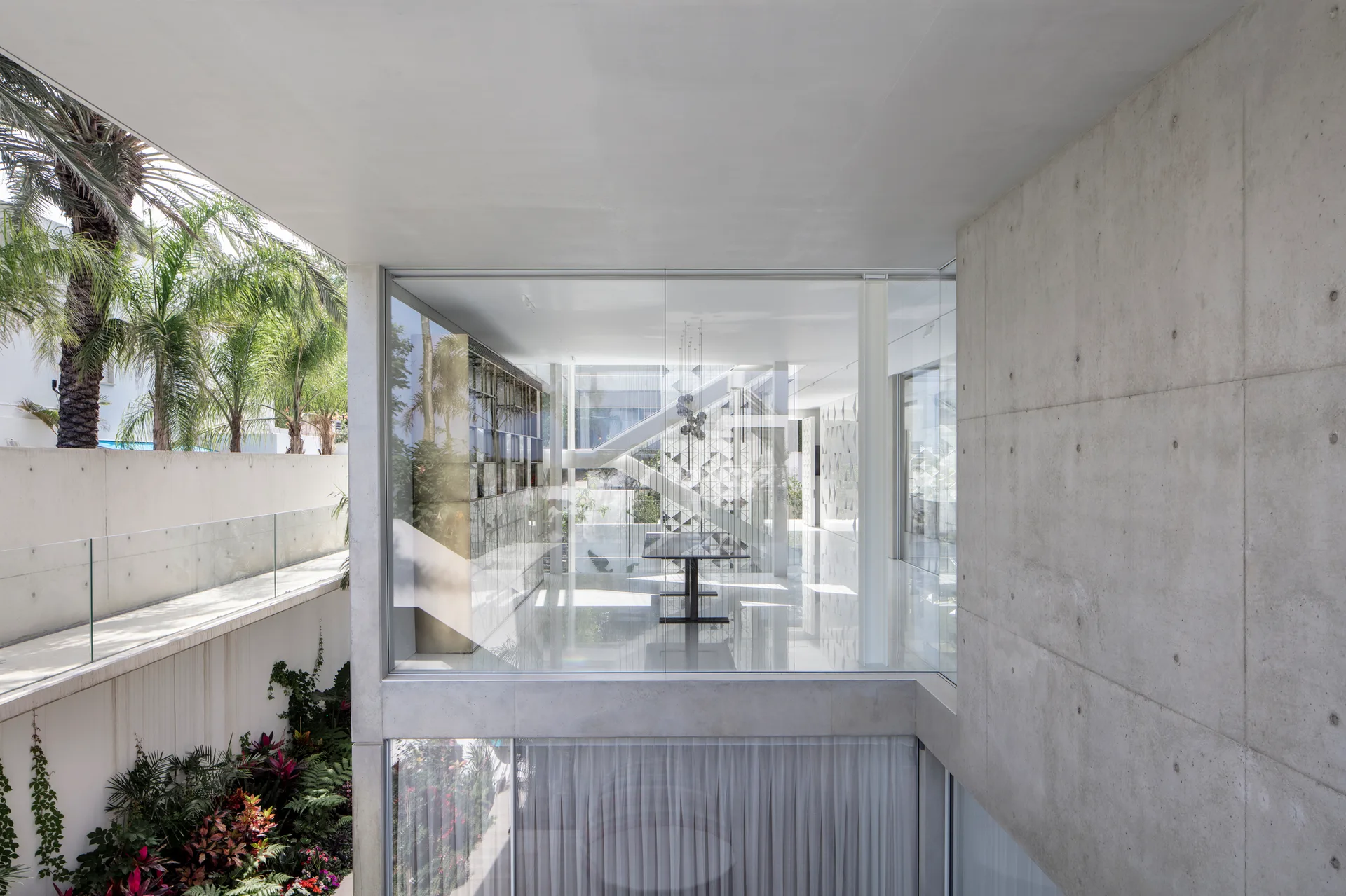 A minimalist dining room features a dark table enclosed by floor-to-ceiling glass walls, set against an exposed concrete wall and a tropical garden.