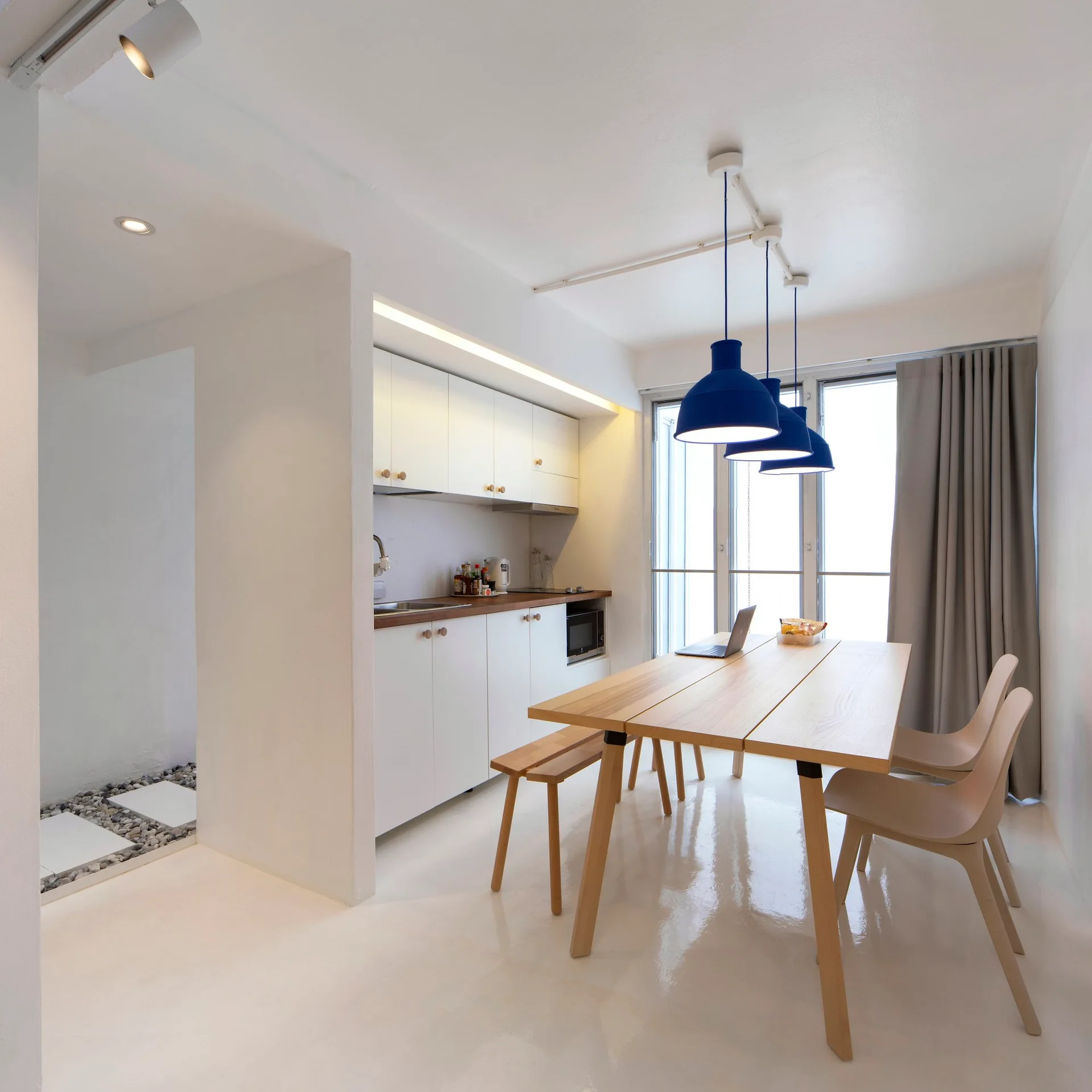 A minimalist open-plan kitchen and dining area features white cabinetry, a light wood dining table with matching seating, and three blue pendant lights.