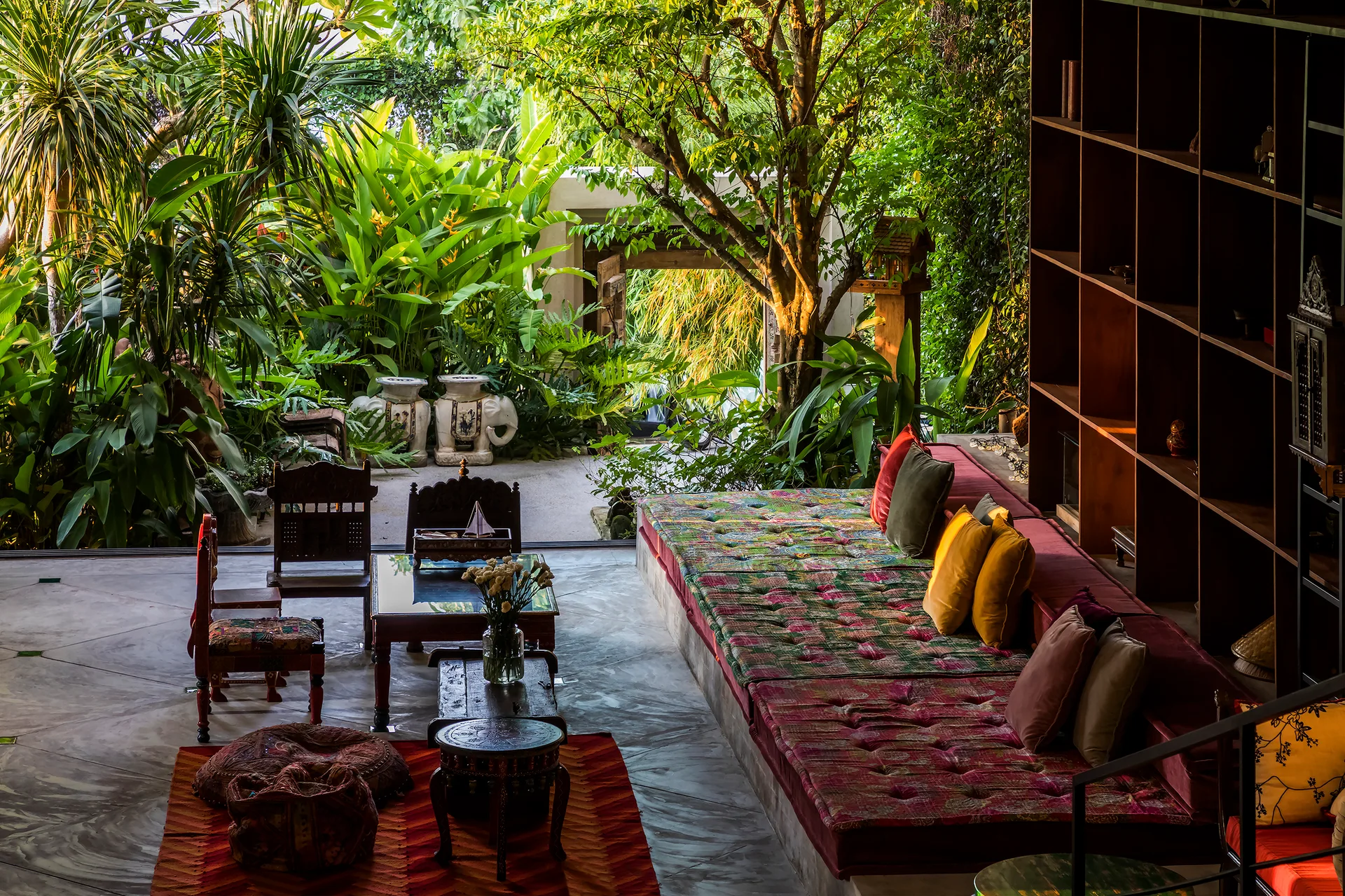 An indoor-outdoor living room features a long built-in daybed with colorful cushions and a large wood bookshelf facing a lush tropical garden.