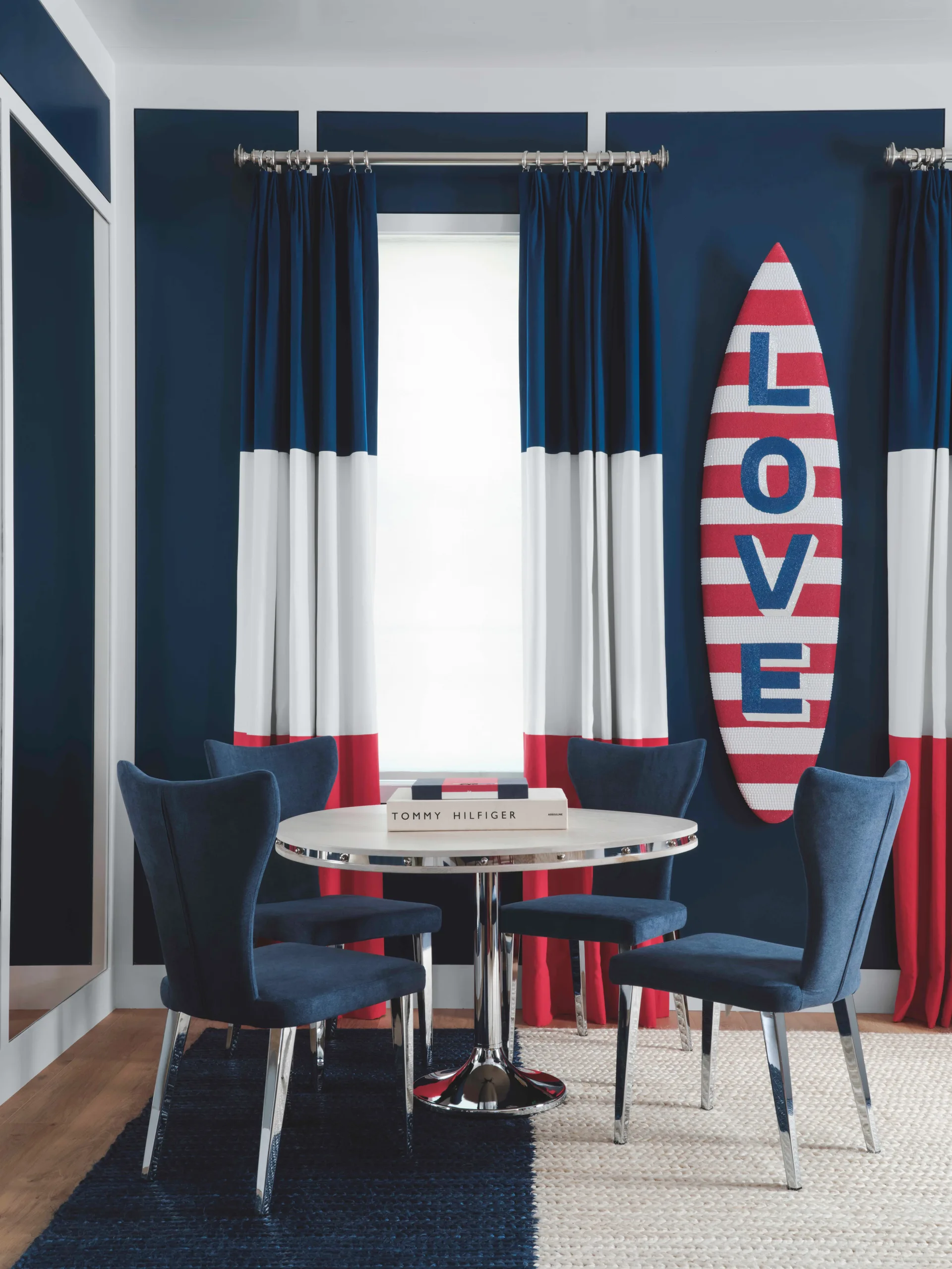 A modern dining set with blue chairs sits against a navy wall decorated with color-blocked curtains and a surfboard.