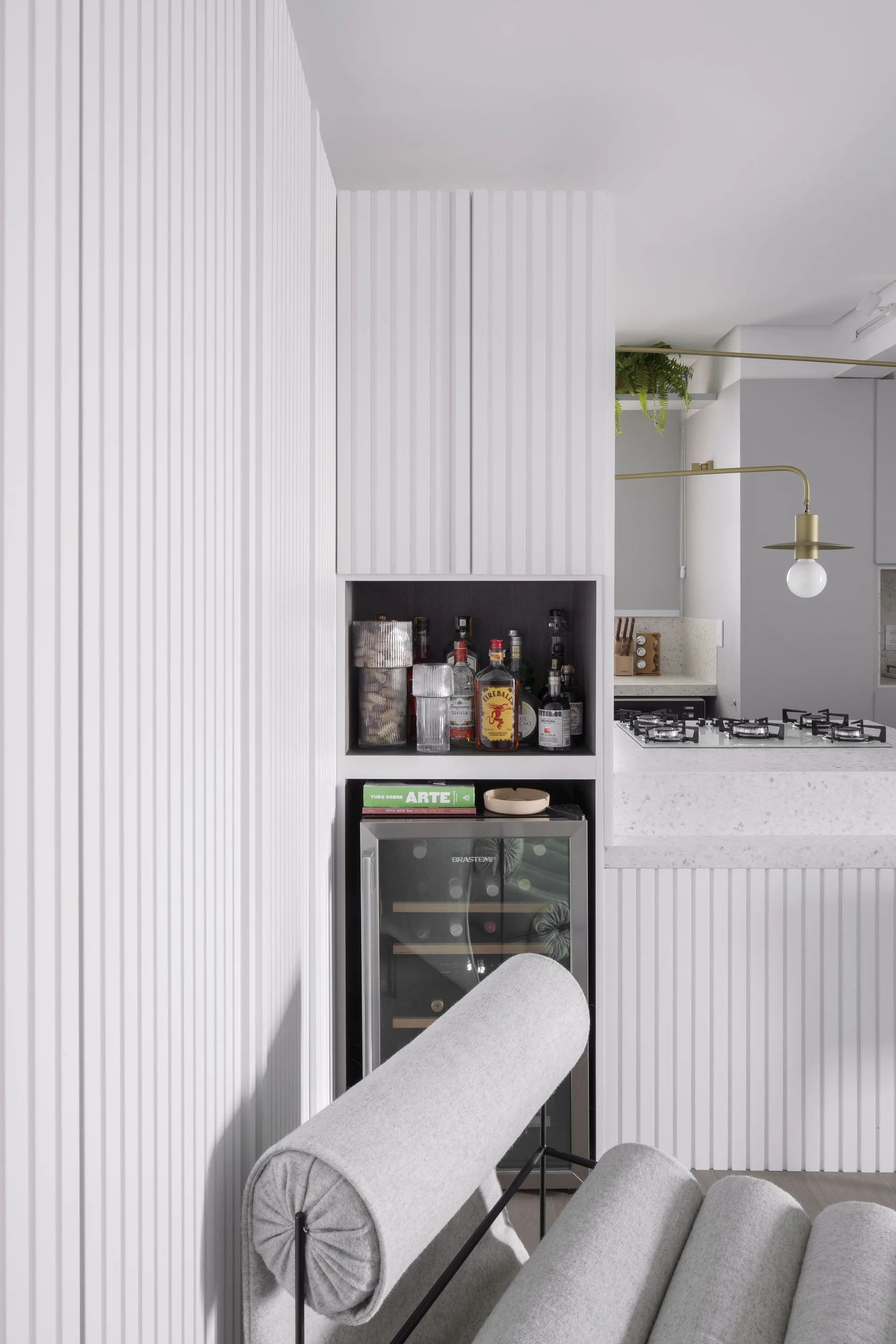 A wine fridge and open bar shelving are integrated into a white slatted cabinet wall, with a grey fabric lounge chair in the foreground.