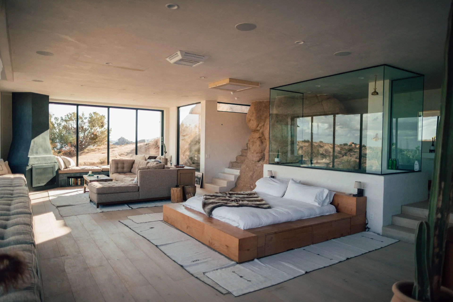 An open-plan desert bedroom features a wooden platform bed, glass-enclosed bathroom, integrated natural rock wall, and a living area with a fireplace.