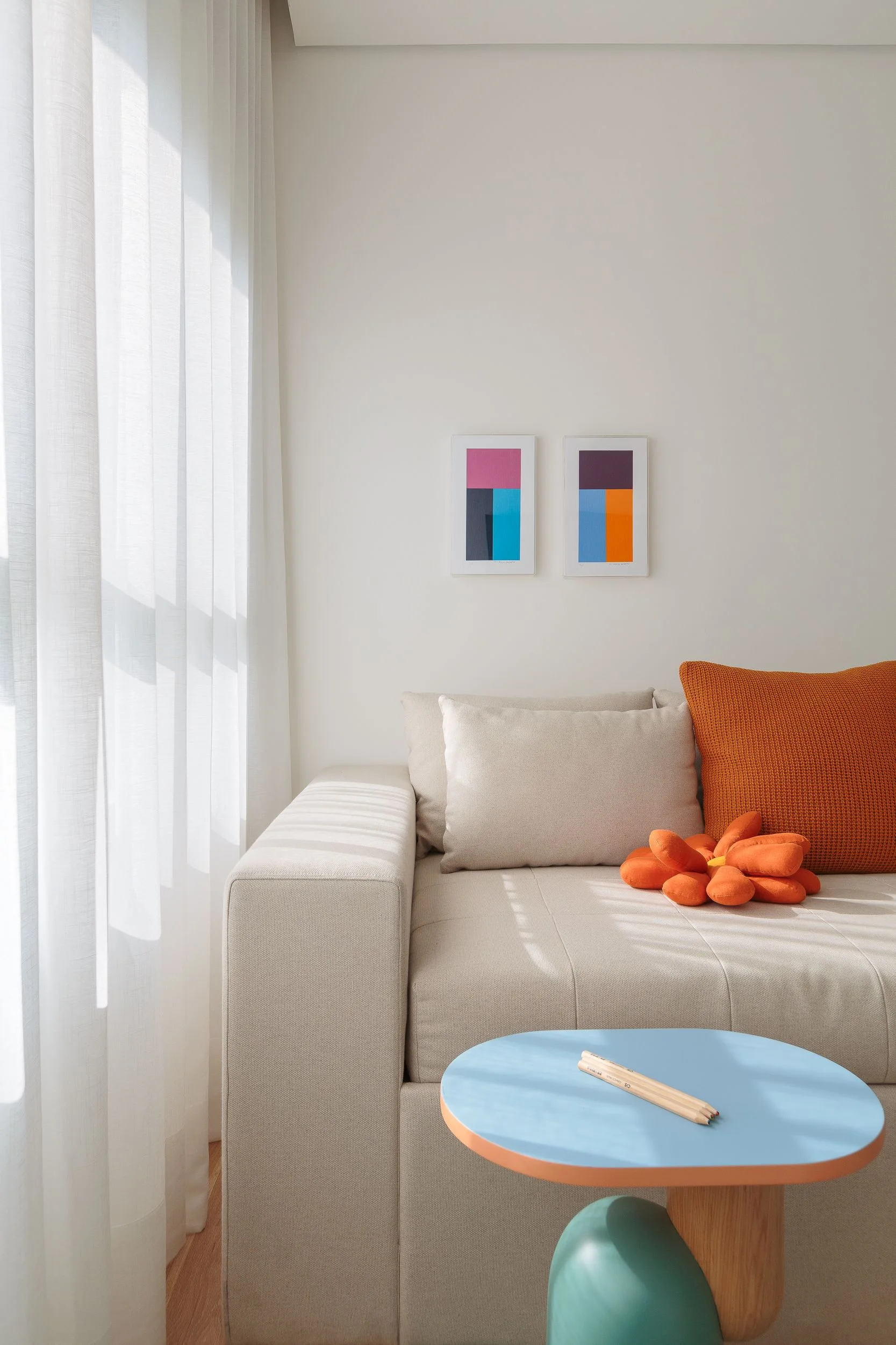 A beige sofa styled with orange pillows sits next to a blue round side table under two colorful geometric art prints on an off-white wall.