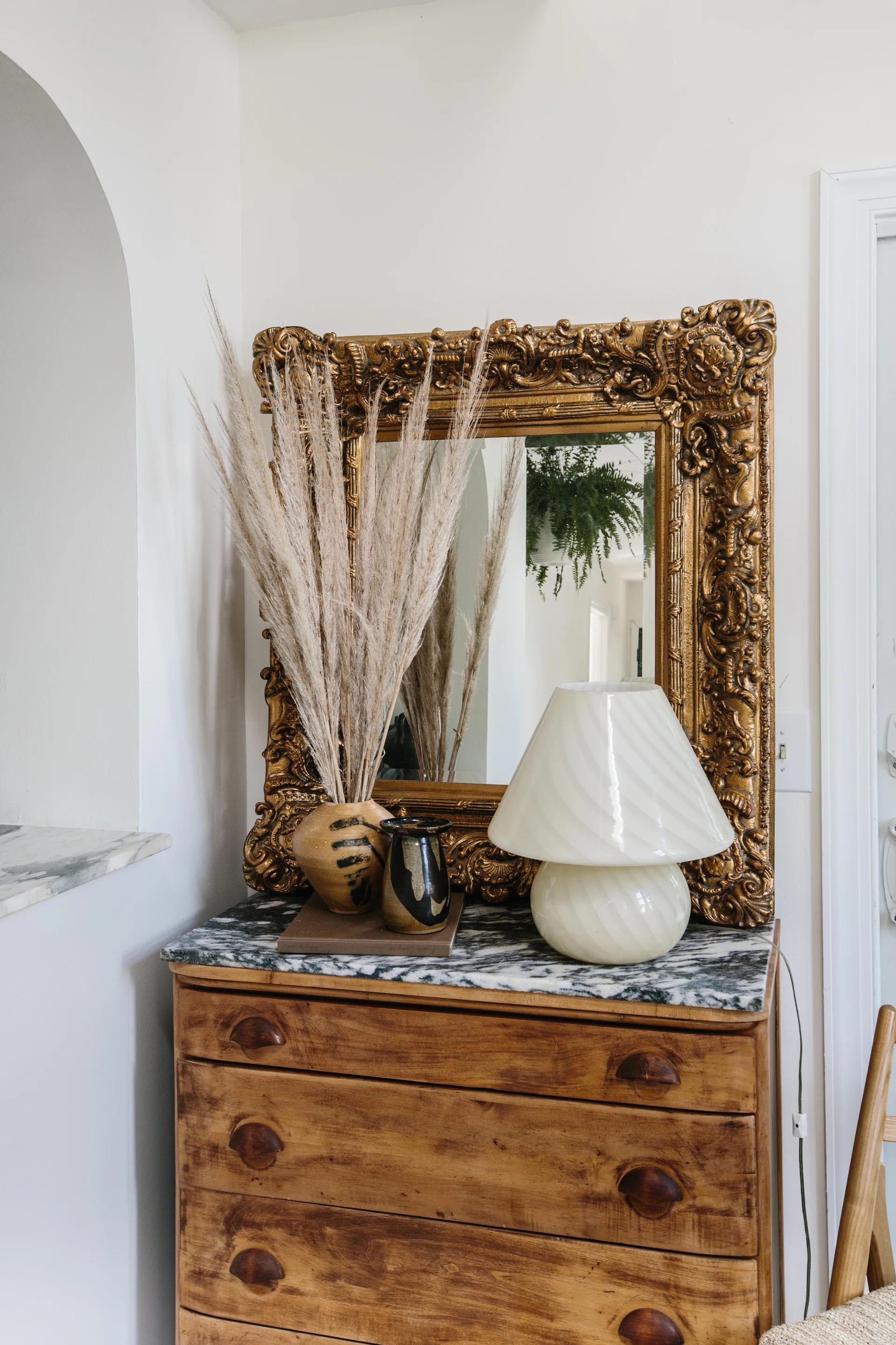 A vintage wooden chest of drawers with a white and grey marble top displaying an ornate gold mirror, a white glass lamp, and dried pampas grass.
