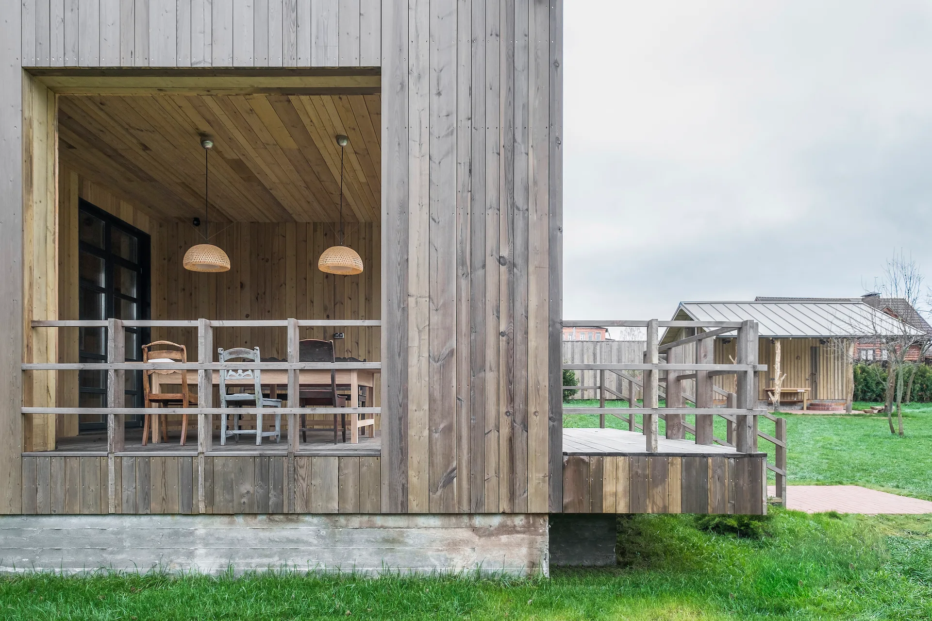 A covered wooden deck features vertical plank siding, a rustic dining table with mismatched chairs, and two woven dome pendant lights.