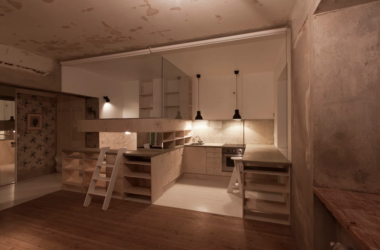 An industrial kitchen area features light plywood cabinetry, concrete countertops, and a raised loft platform with a clear glass safety panel.