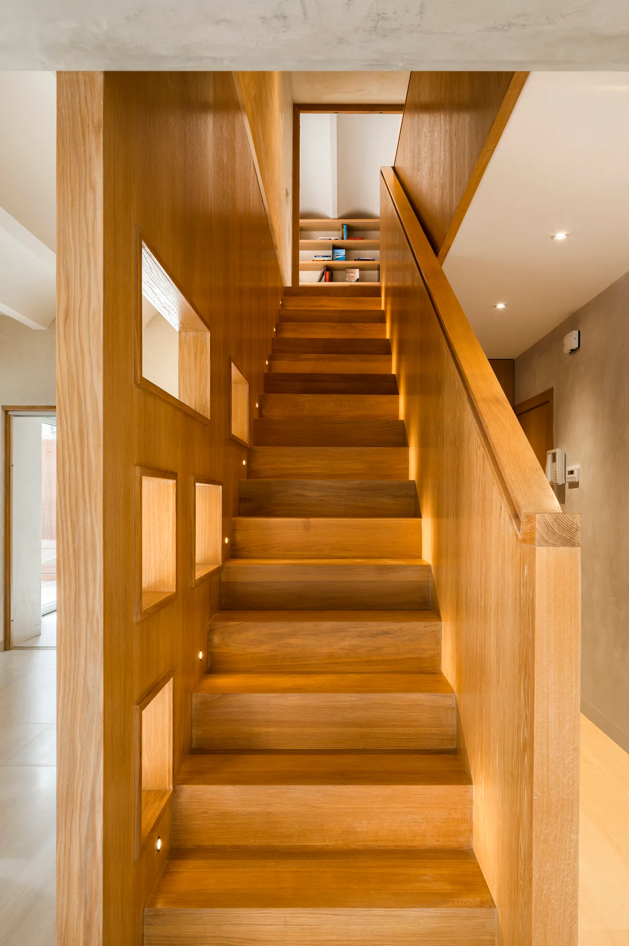 A modern wood-paneled staircase features integrated warm accent lights along the stairs and square geometric cutouts in the side wall.