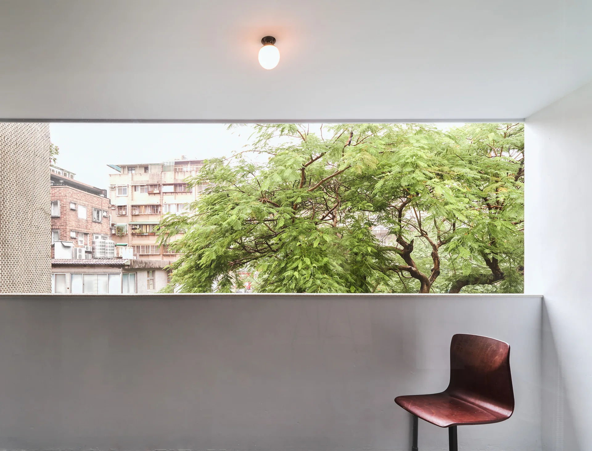 A minimalist white balcony room features a dark wood chair and a wide rectangular window looking out onto green leafy trees.