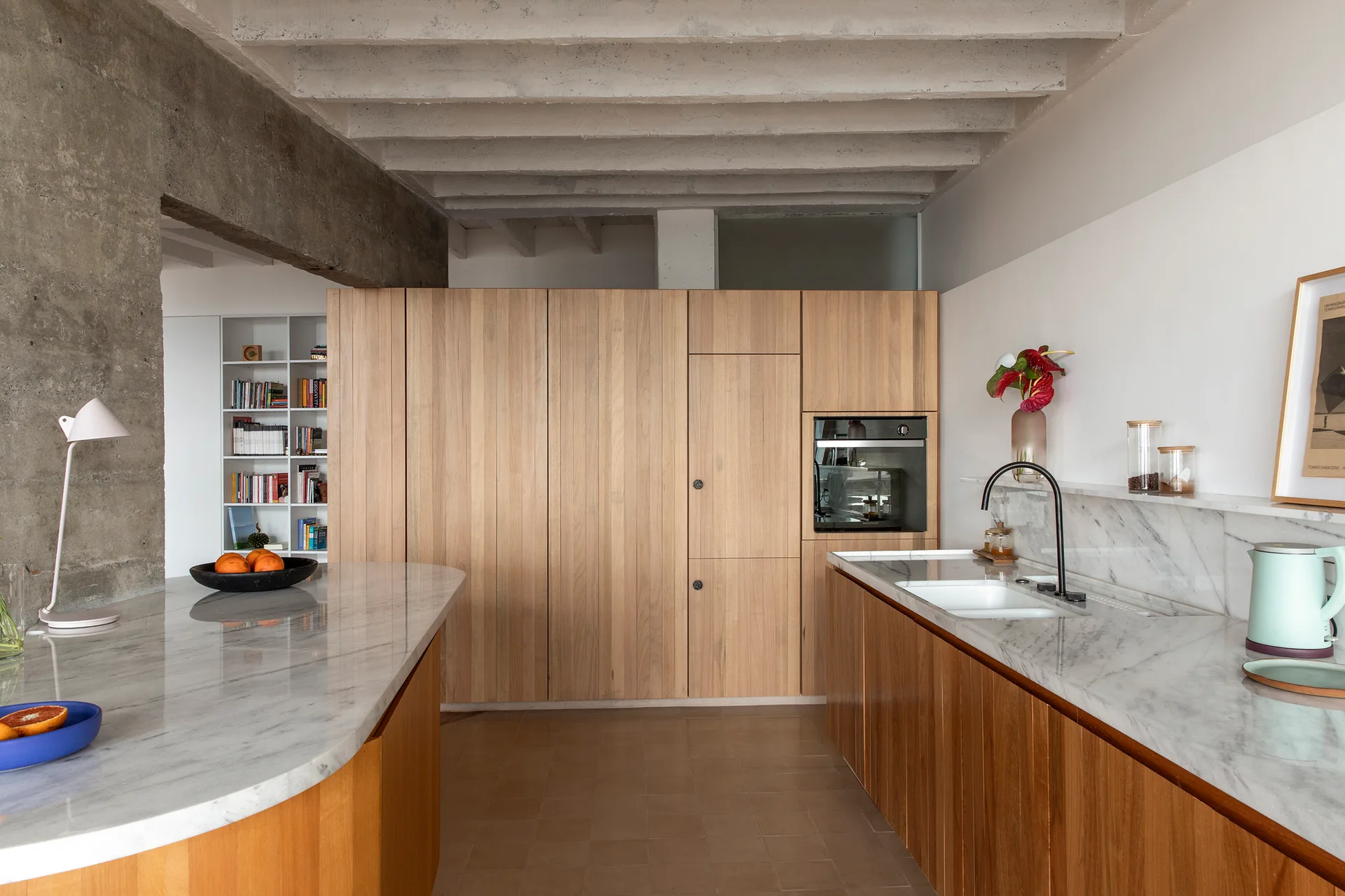 A modern kitchen featuring light wood flat-panel cabinets, marble countertops, an island, and exposed raw concrete ceiling beams.