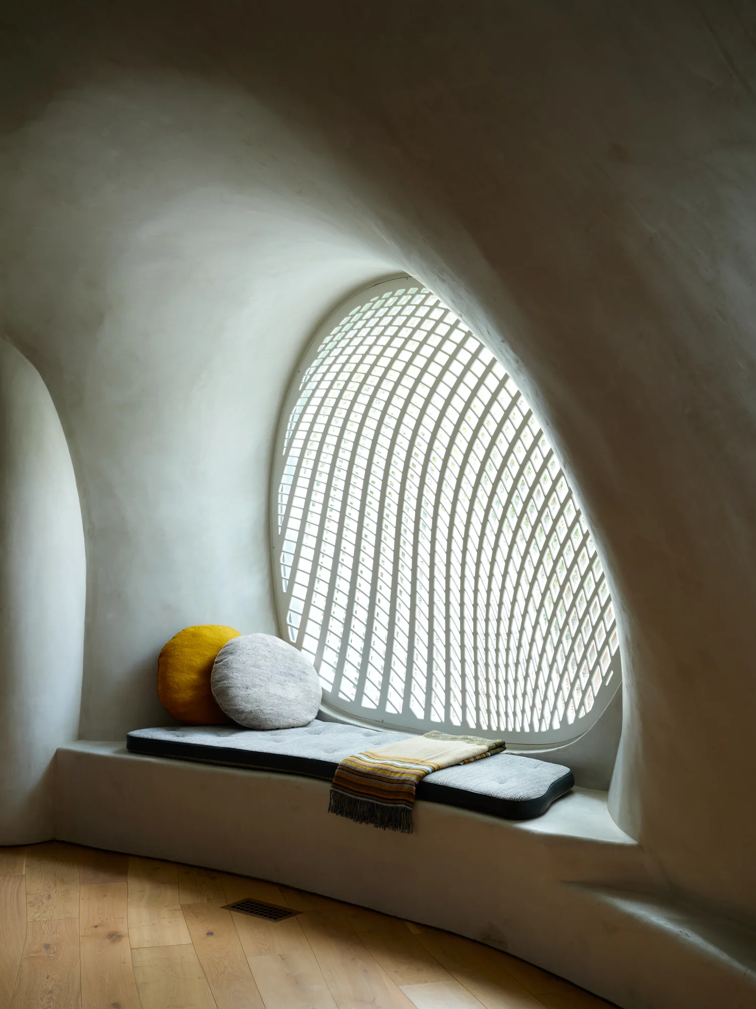 A built-in window seat with grey cushions and round pillows sits inside a curved white plaster niche under a geometric patterned window.
