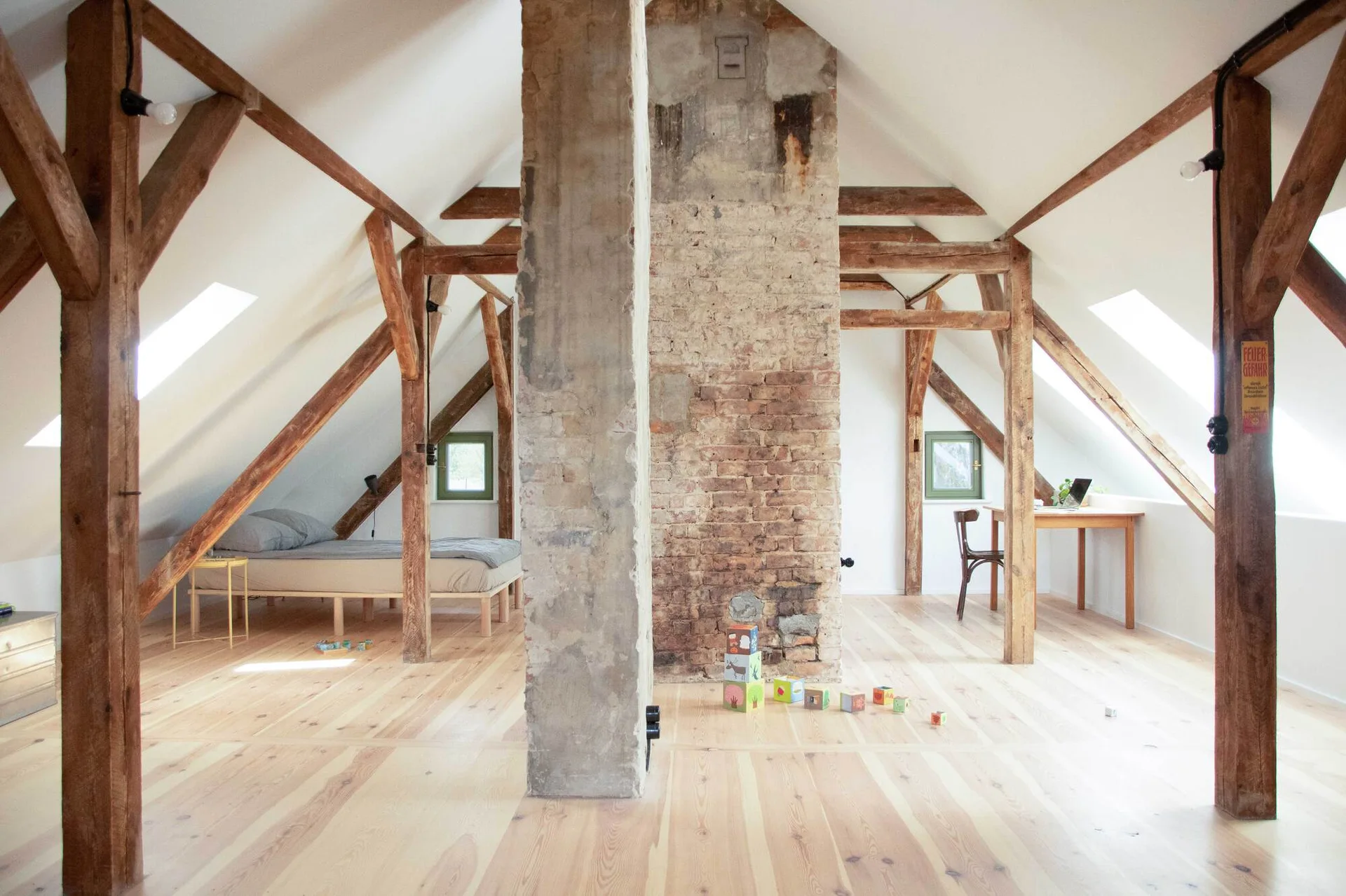 An attic bedroom with exposed timber beams, light wood flooring, skylights, and a central weathered brick chimney.