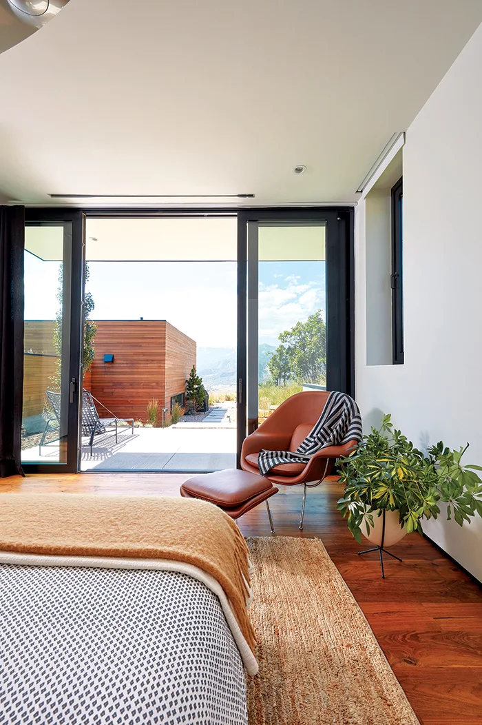 A modern bedroom features a bed with a mustard throw, a cognac leather lounge chair, and large sliding glass doors opening to a wood-sided patio.