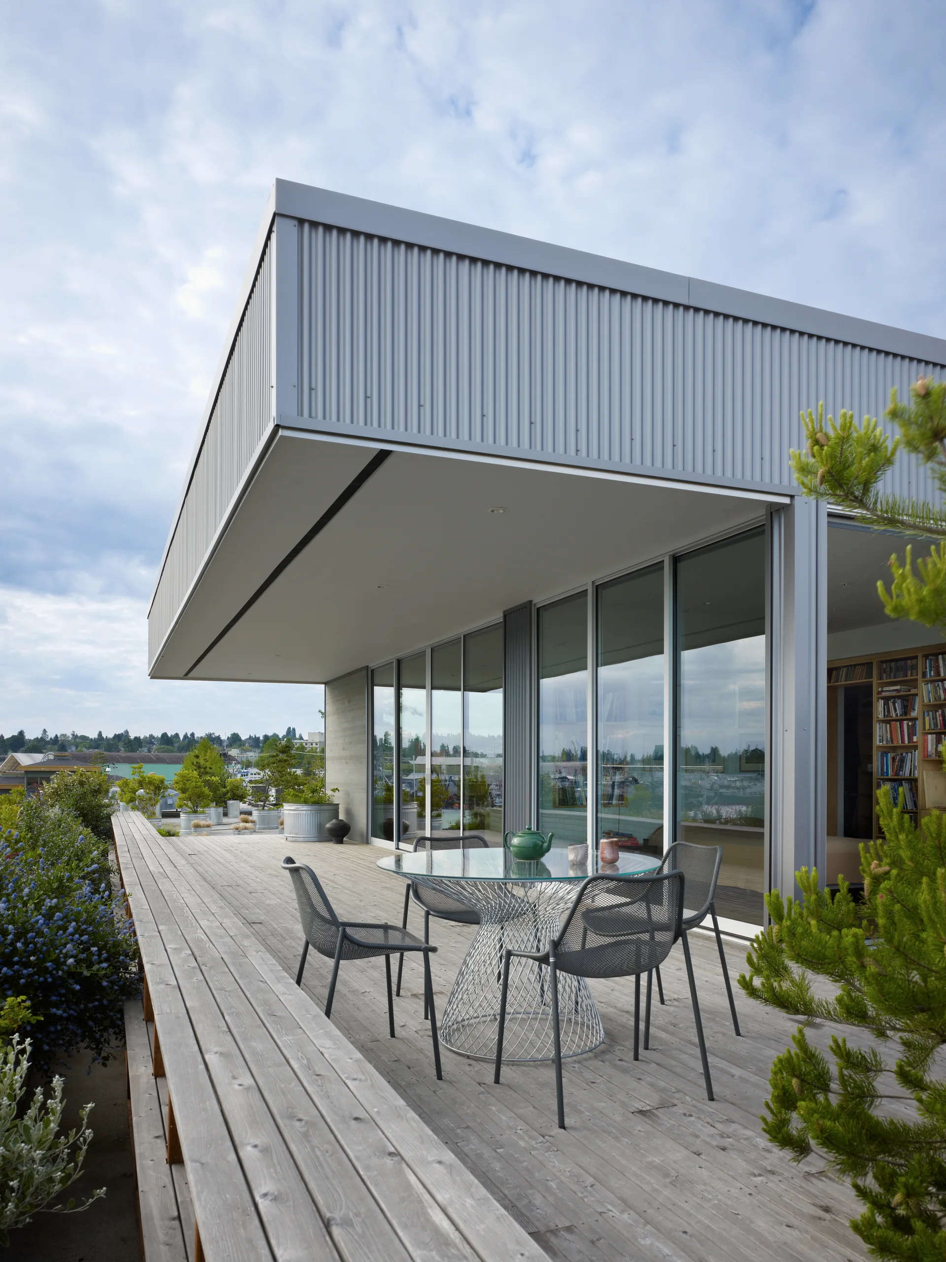 A modern outdoor wooden deck features a circular glass-top table with four grey wire-mesh chairs set against corrugated metal siding and glass walls.