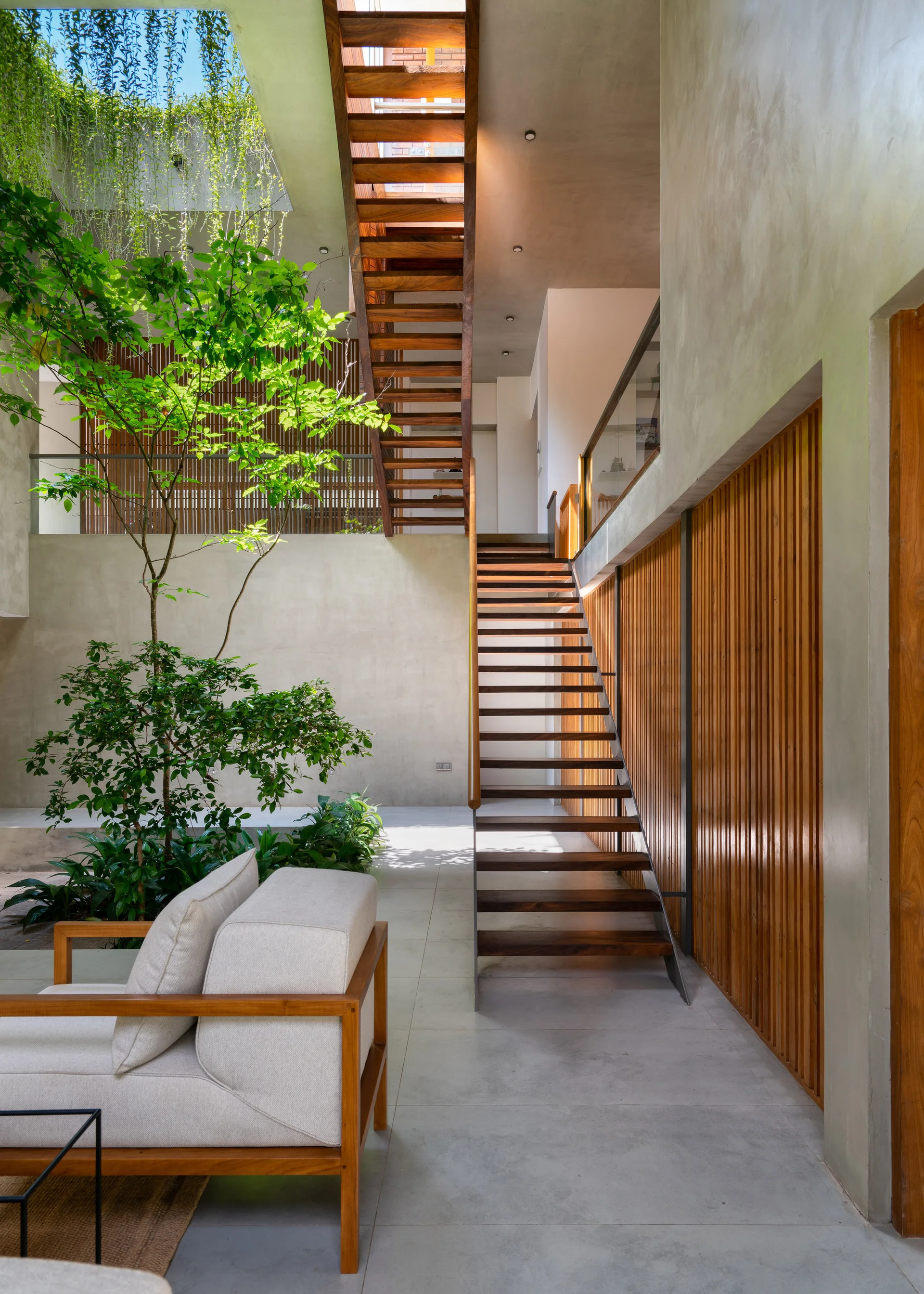 A modern indoor courtyard with a central open-tread wooden staircase, a green indoor tree, smooth concrete walls, and vertical wood slat paneling.