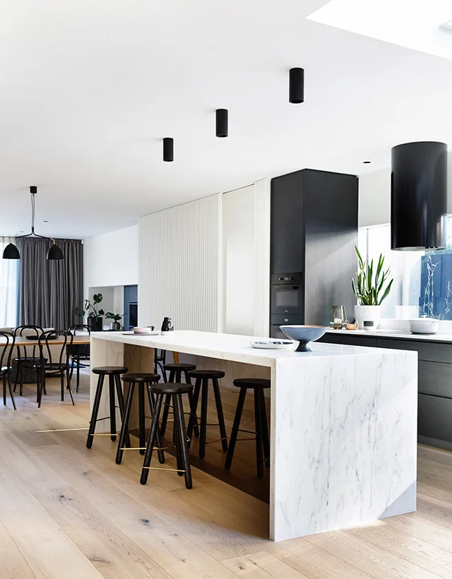 A modern open-plan kitchen featuring a white marble waterfall island, dark wood bar stools, ribbed wall paneling, and black cylindrical ceiling lights.