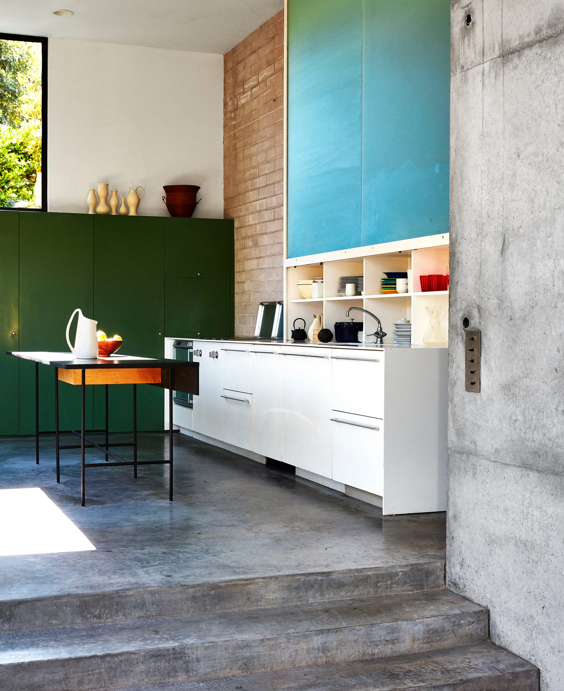 A modern kitchen features color-blocked blue and green cabinets, an exposed brick wall, white base cabinets, and a polished concrete floor.