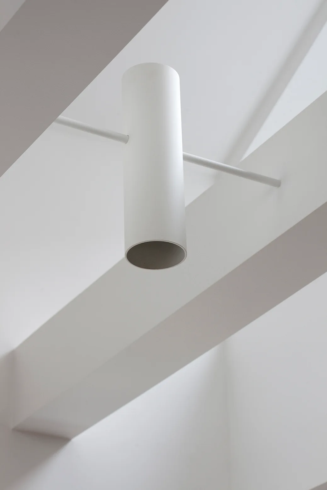 A matte white cylindrical pendant light hangs from a thin rod between two white architectural ceiling beams.