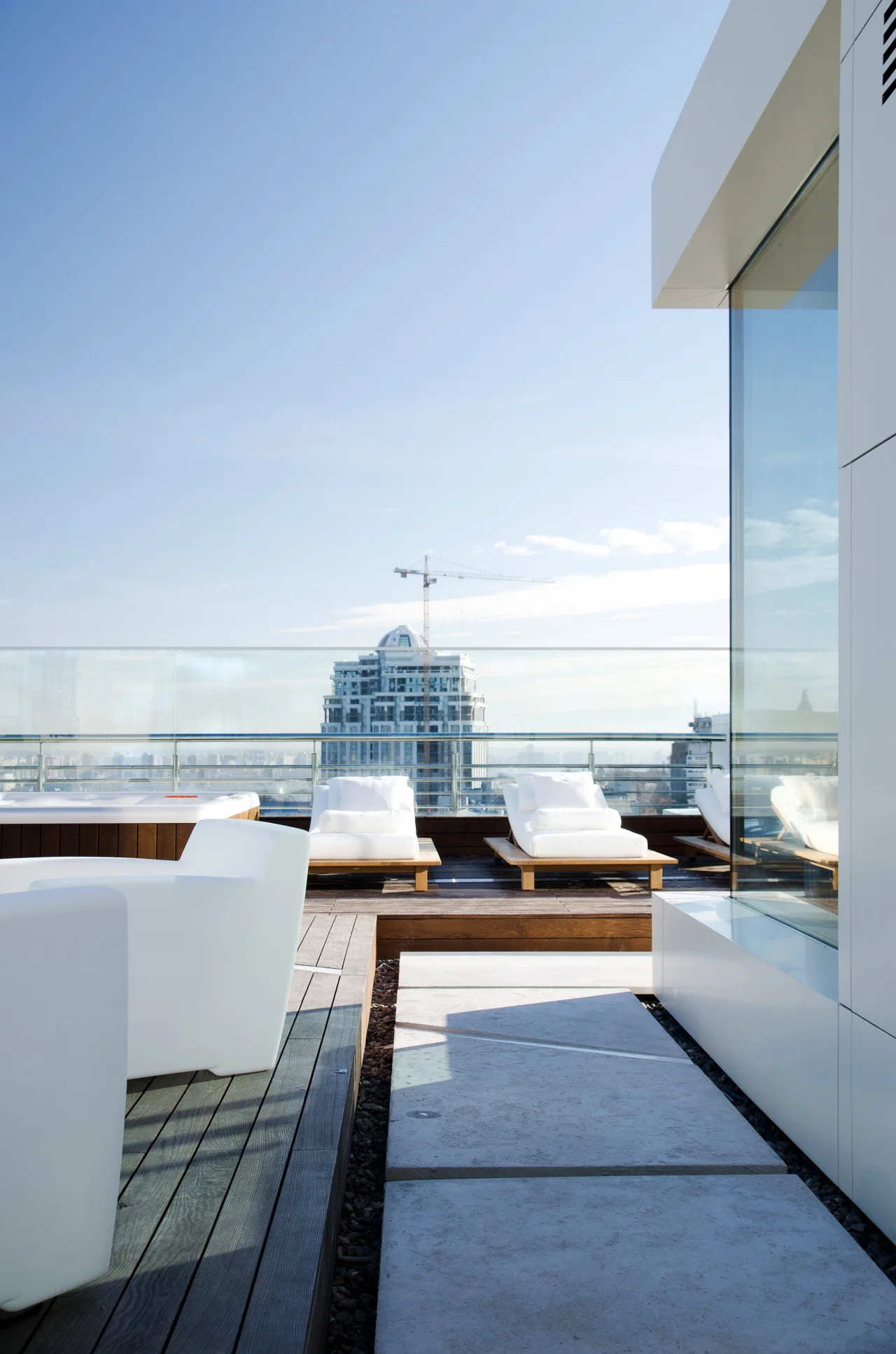 White lounge chairs and armchairs rest on a modern wooden rooftop deck featuring a hot tub and a clear glass railing overlooking the city.