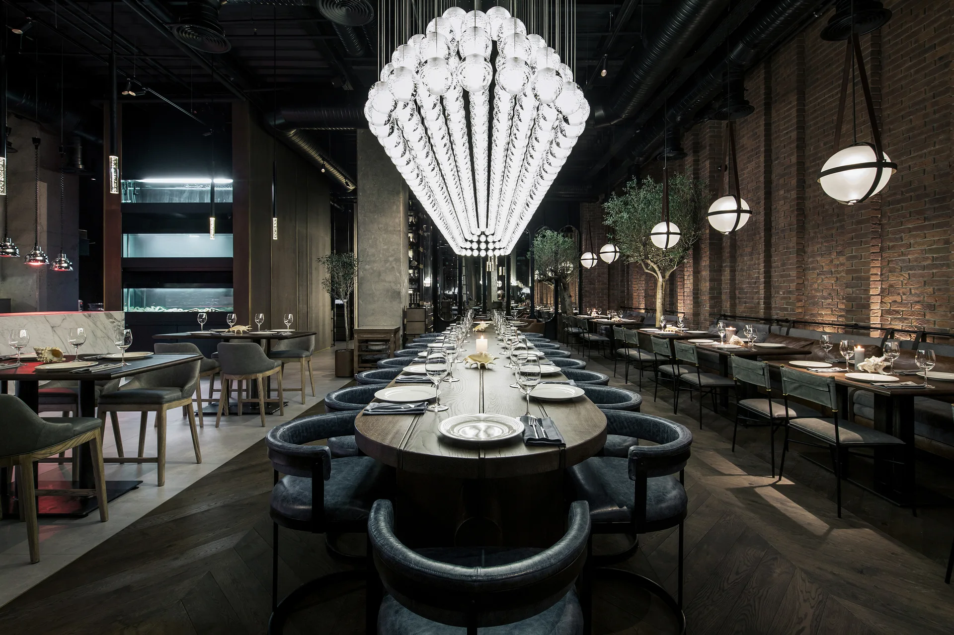 A spacious dining room features a long wooden table beneath a massive cascading sphere chandelier, bordered by exposed brick walls and dark seating.