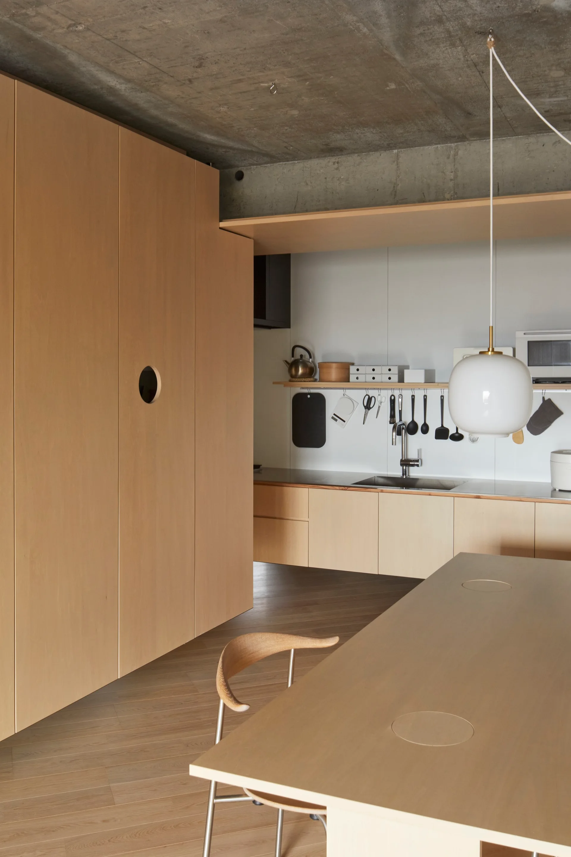 A minimalist kitchen features light wood cabinetry with a circular cutout handle, an exposed concrete ceiling, and a matching wooden table.