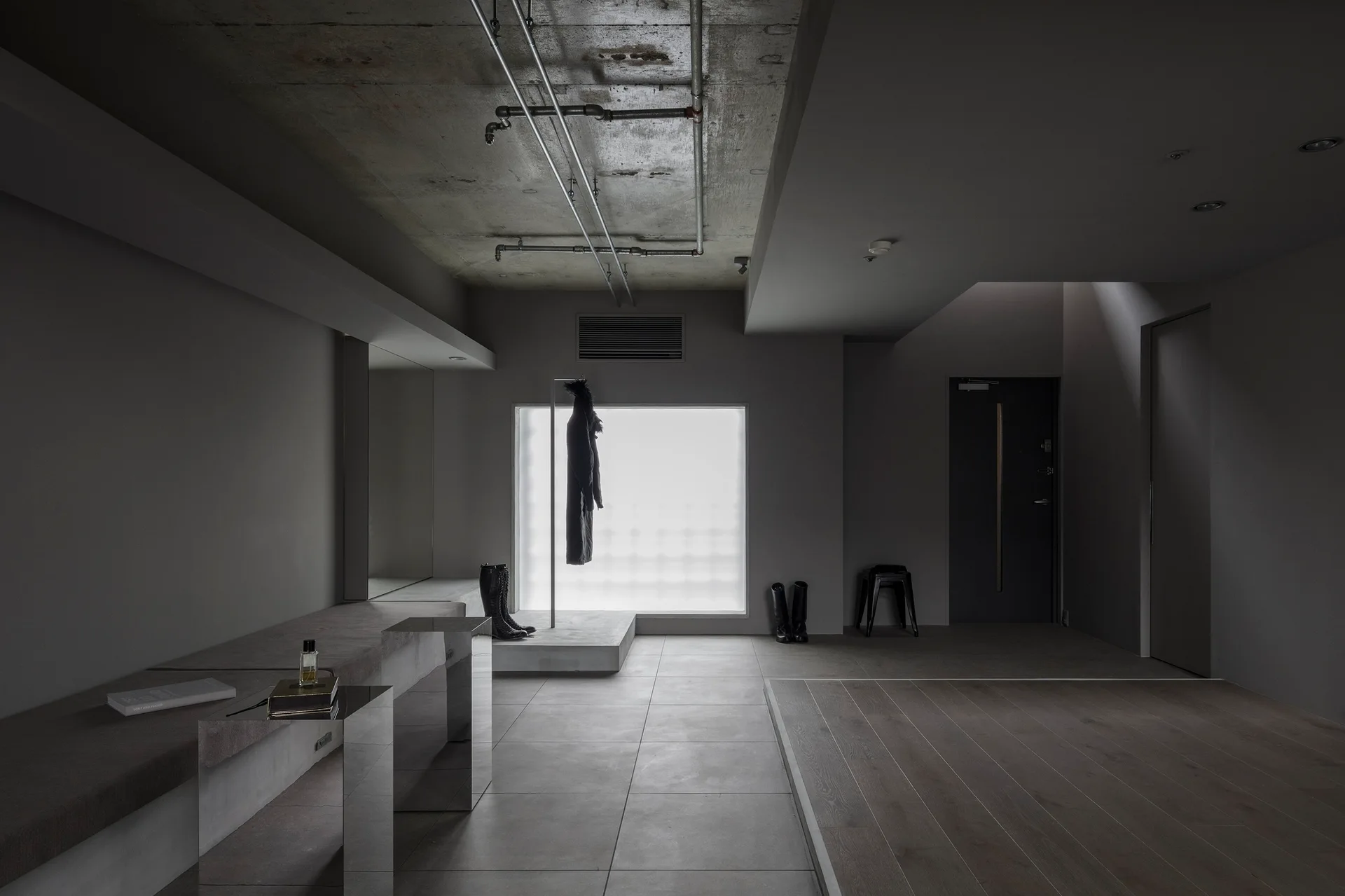 A gray minimalist entryway features a raw concrete ceiling with exposed pipes, a long concrete bench with a mirrored side, and a large frosted window.
