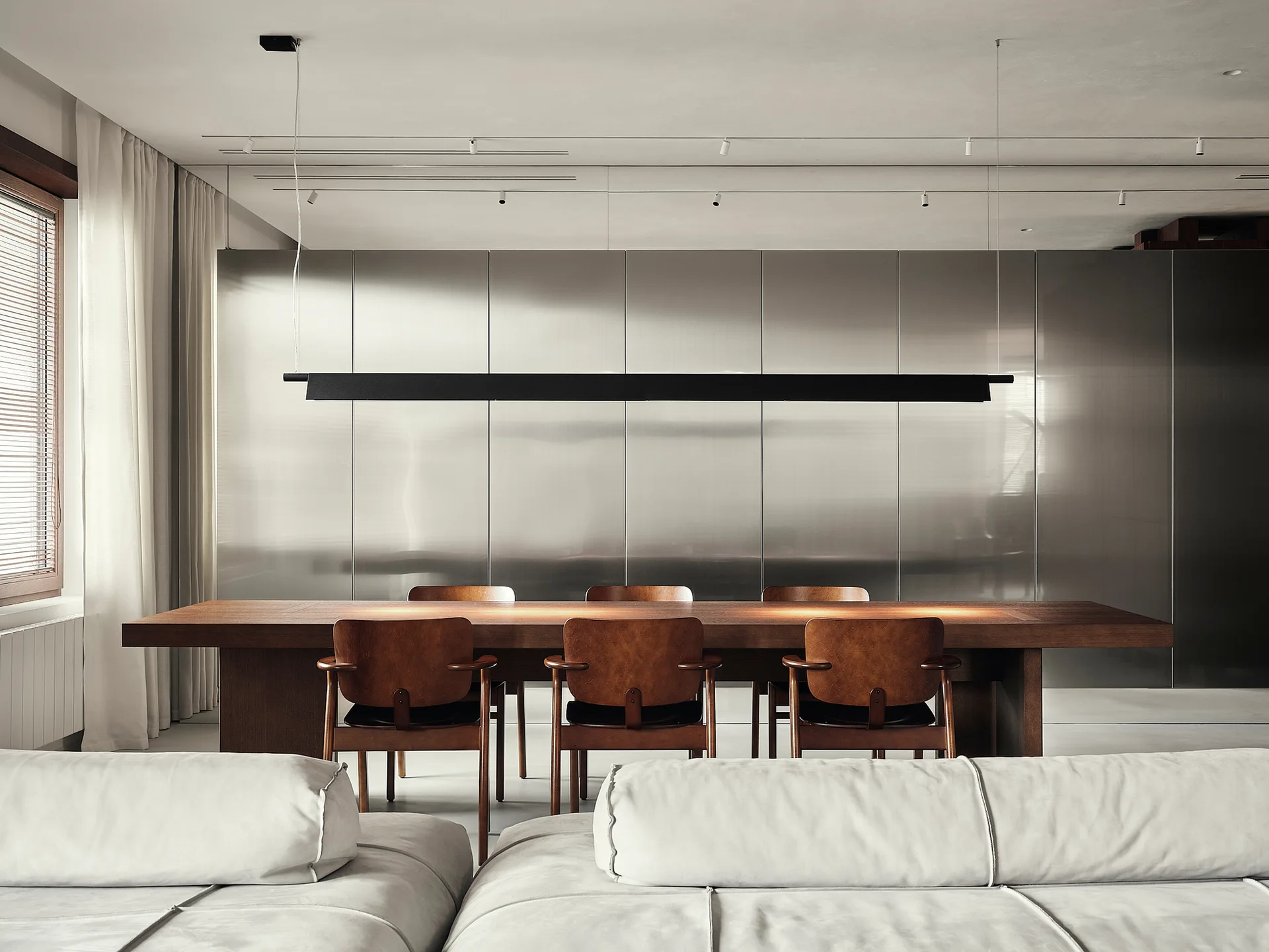 A large wooden dining table and matching chairs are arranged in front of a reflective metallic wall under a long black linear pendant light.