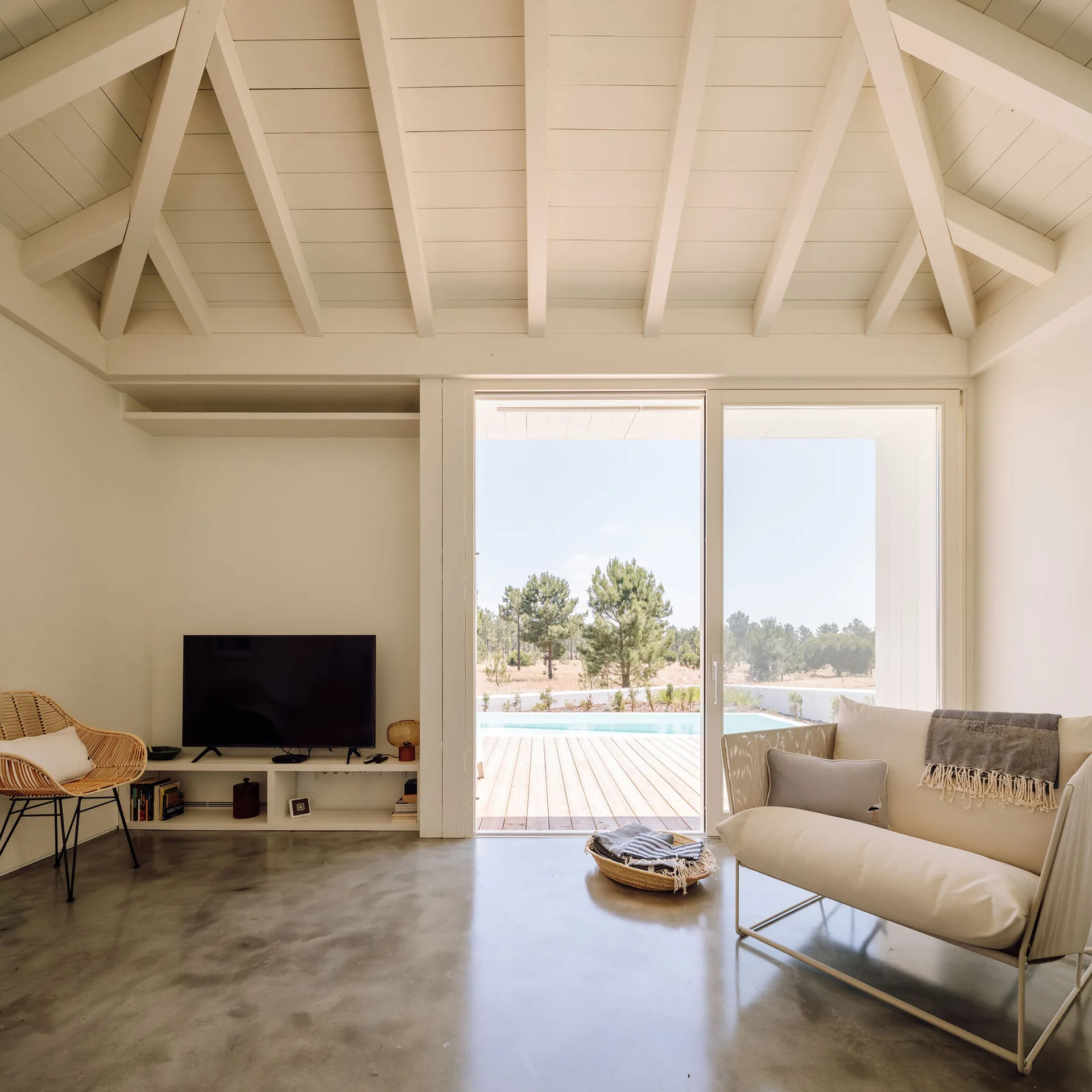 A living room features a vaulted wood-beam ceiling, polished concrete floor, a white sofa, and sliding glass doors opening to an outdoor pool.