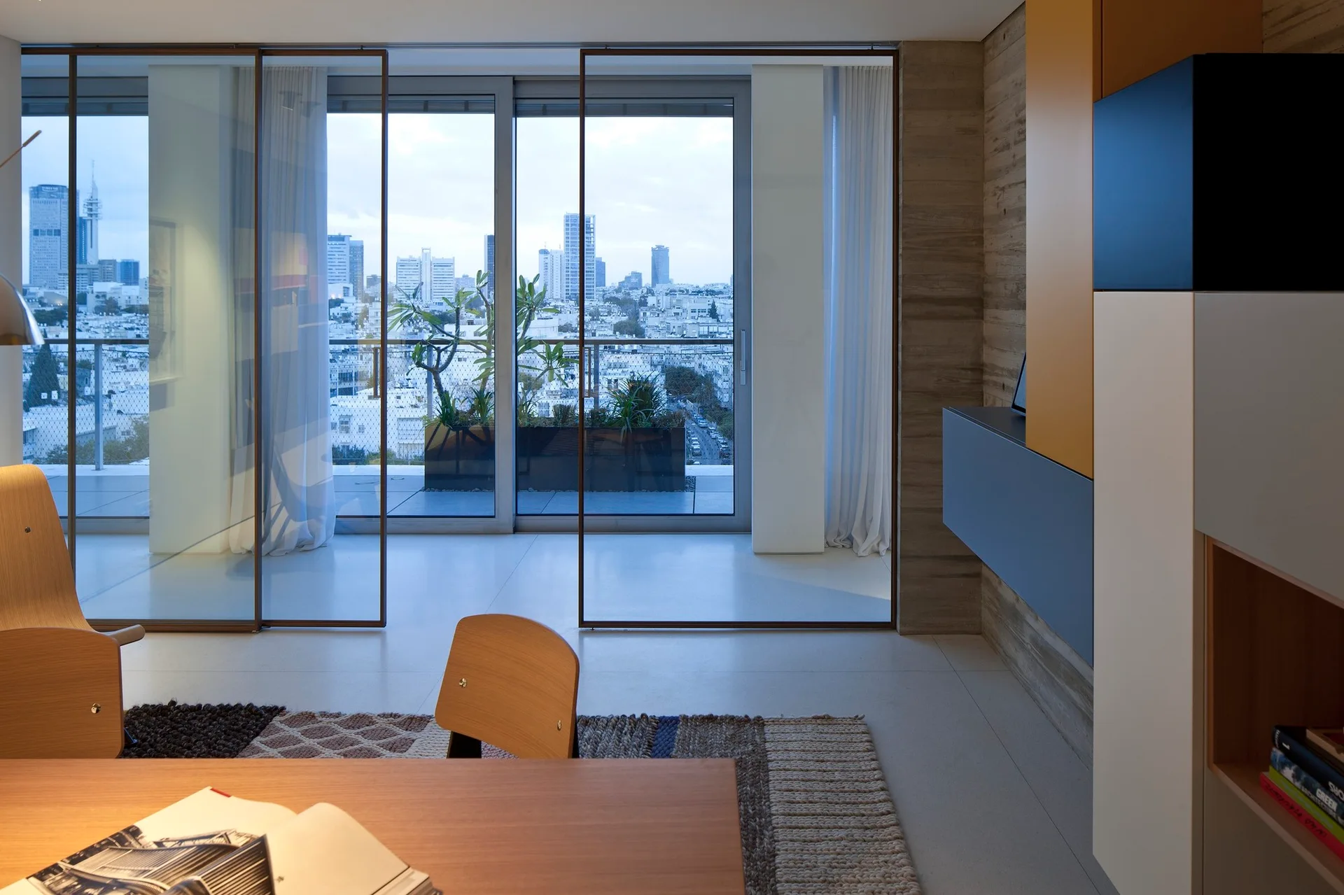 A modern workspace with a wooden desk and chairs next to thin-framed sliding glass doors opening to a city balcony.