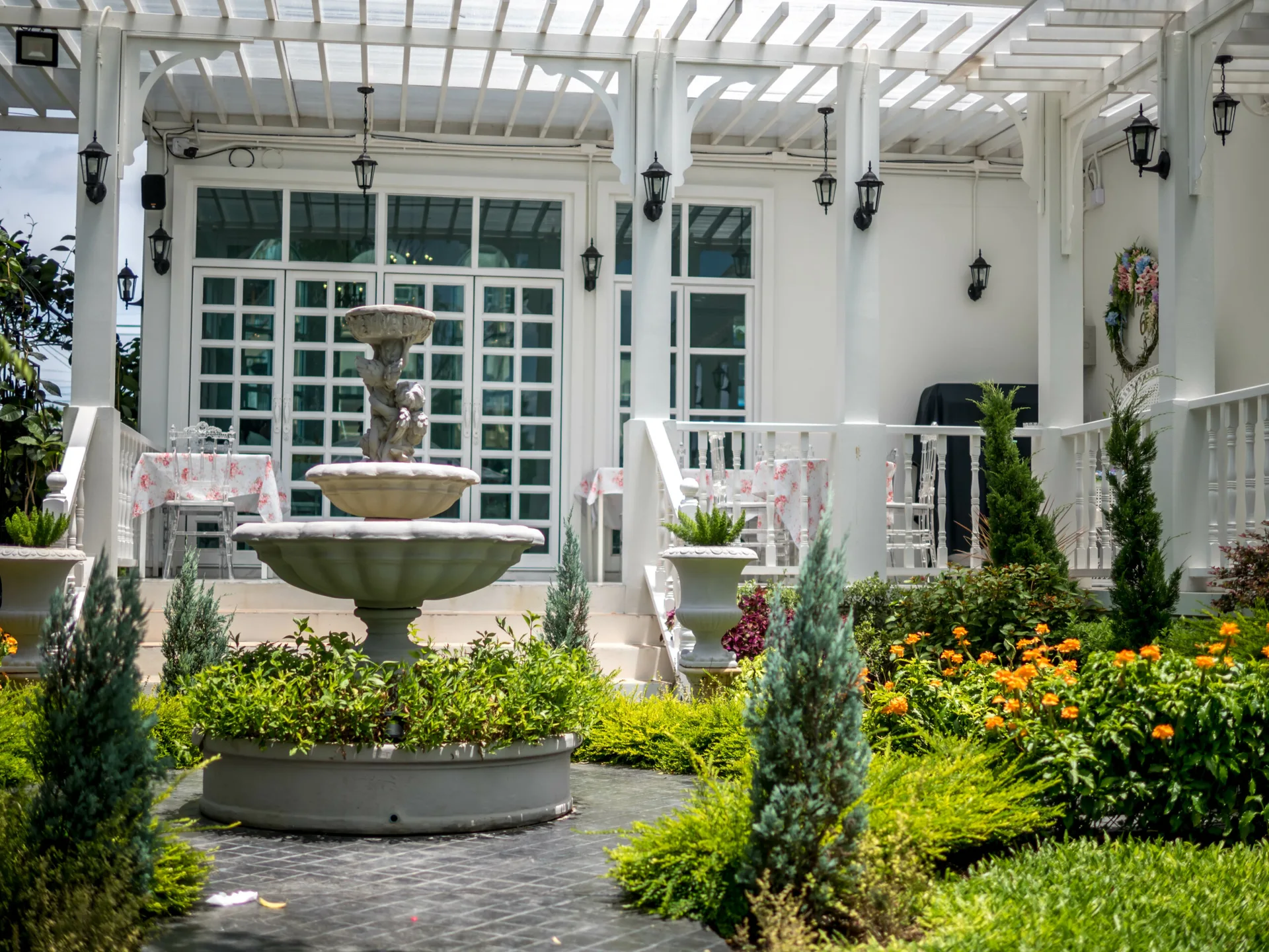A tiered stone fountain stands in a green courtyard in front of a white wooden pergola deck with outdoor tables and black wall lanterns.
