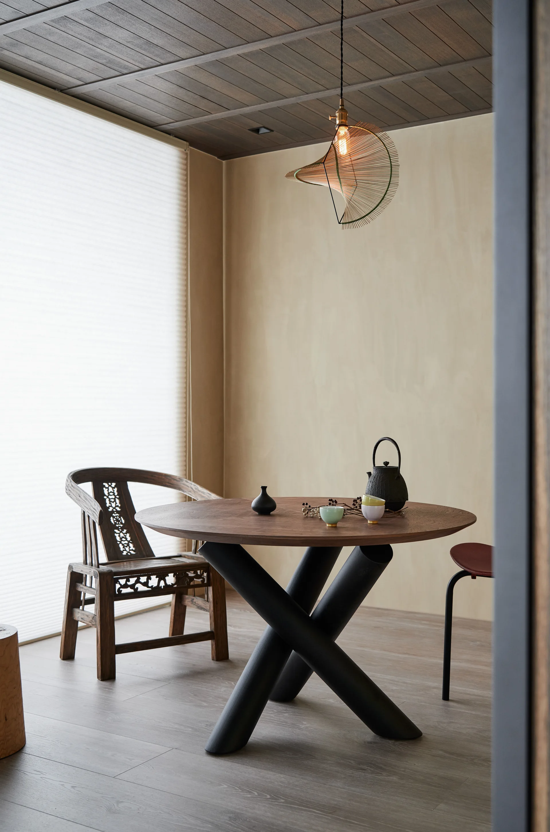 A round wooden dining table with crossed black legs paired with an antique carved wood chair under a dark wood plank ceiling.