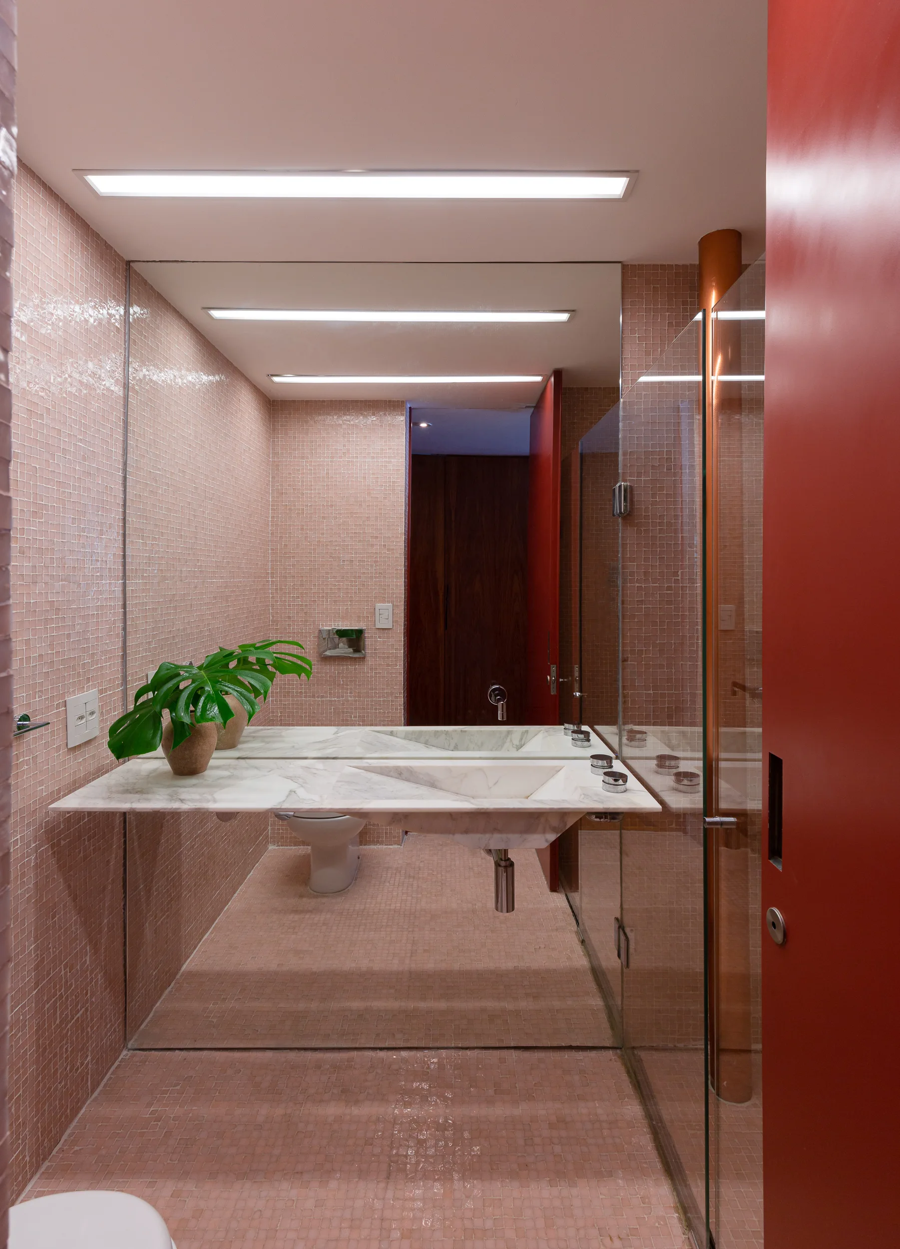A minimalist bathroom features pink mosaic tile walls and flooring, a white marble floating vanity with an integrated sink, and a bold red door.