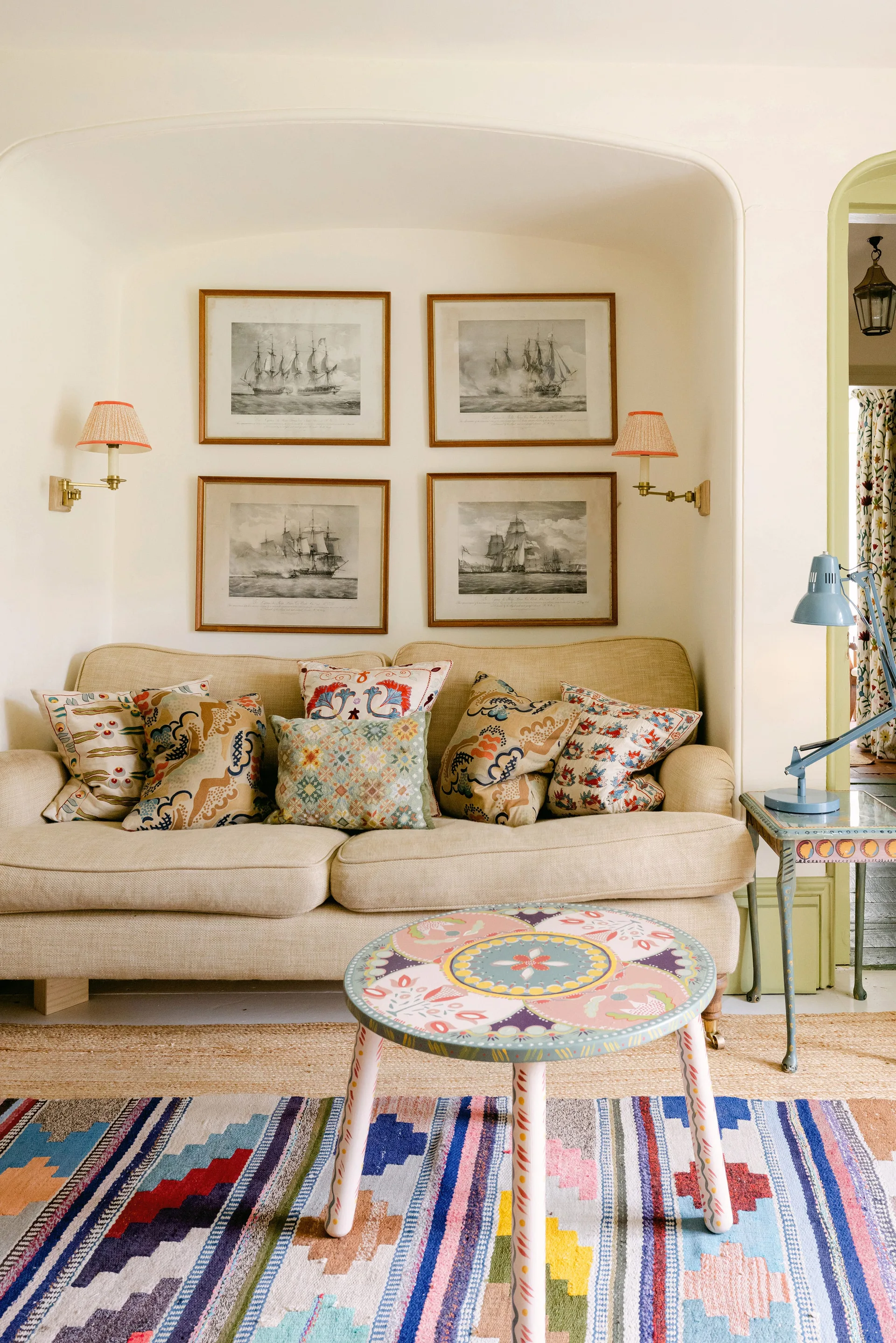A cream-colored sofa sits inside an arched wall alcove under four framed nautical prints, flanked by sconces and a patterned coffee table.