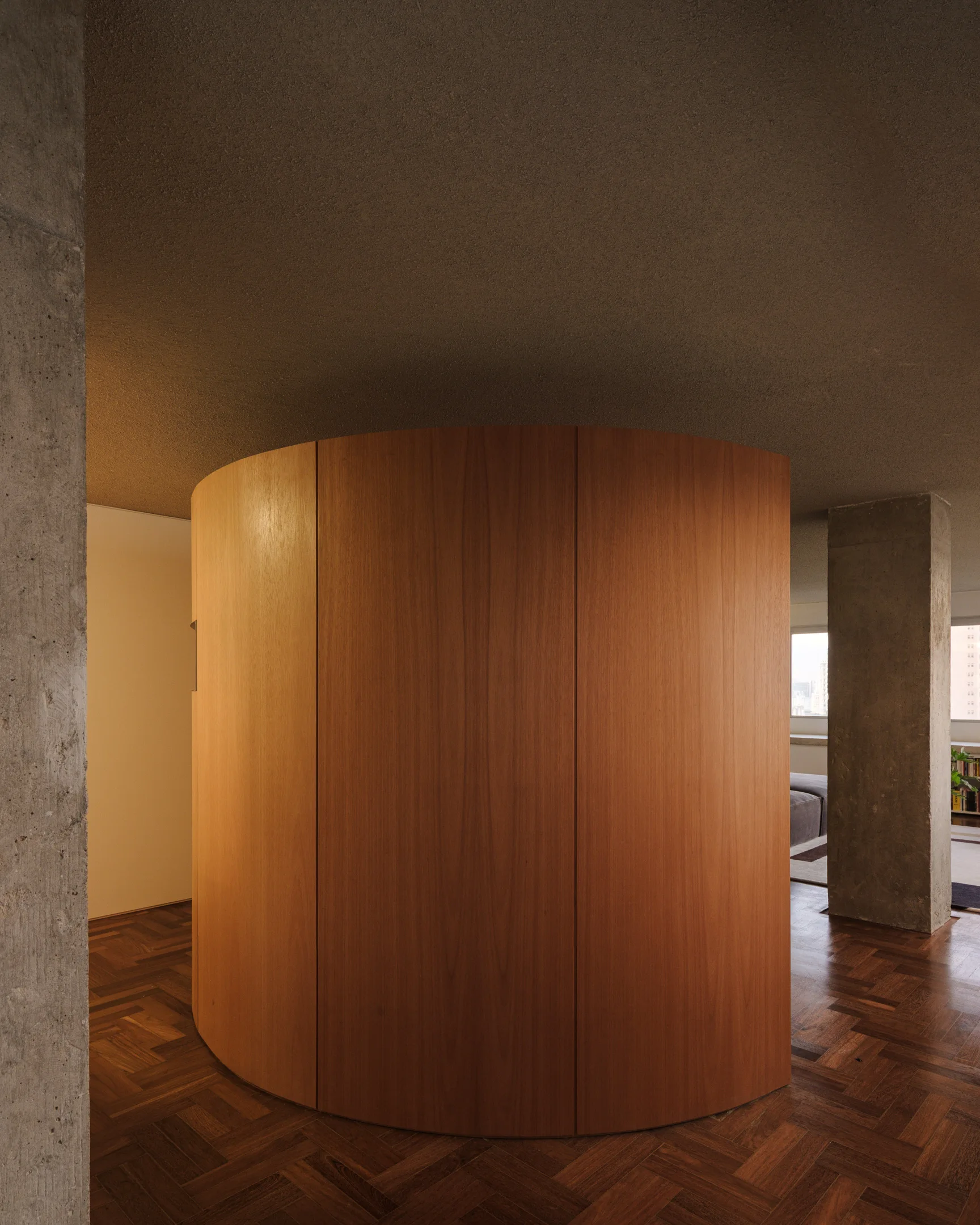 A curved wood panel partition stands between raw concrete columns on a dark herringbone parquet floor.