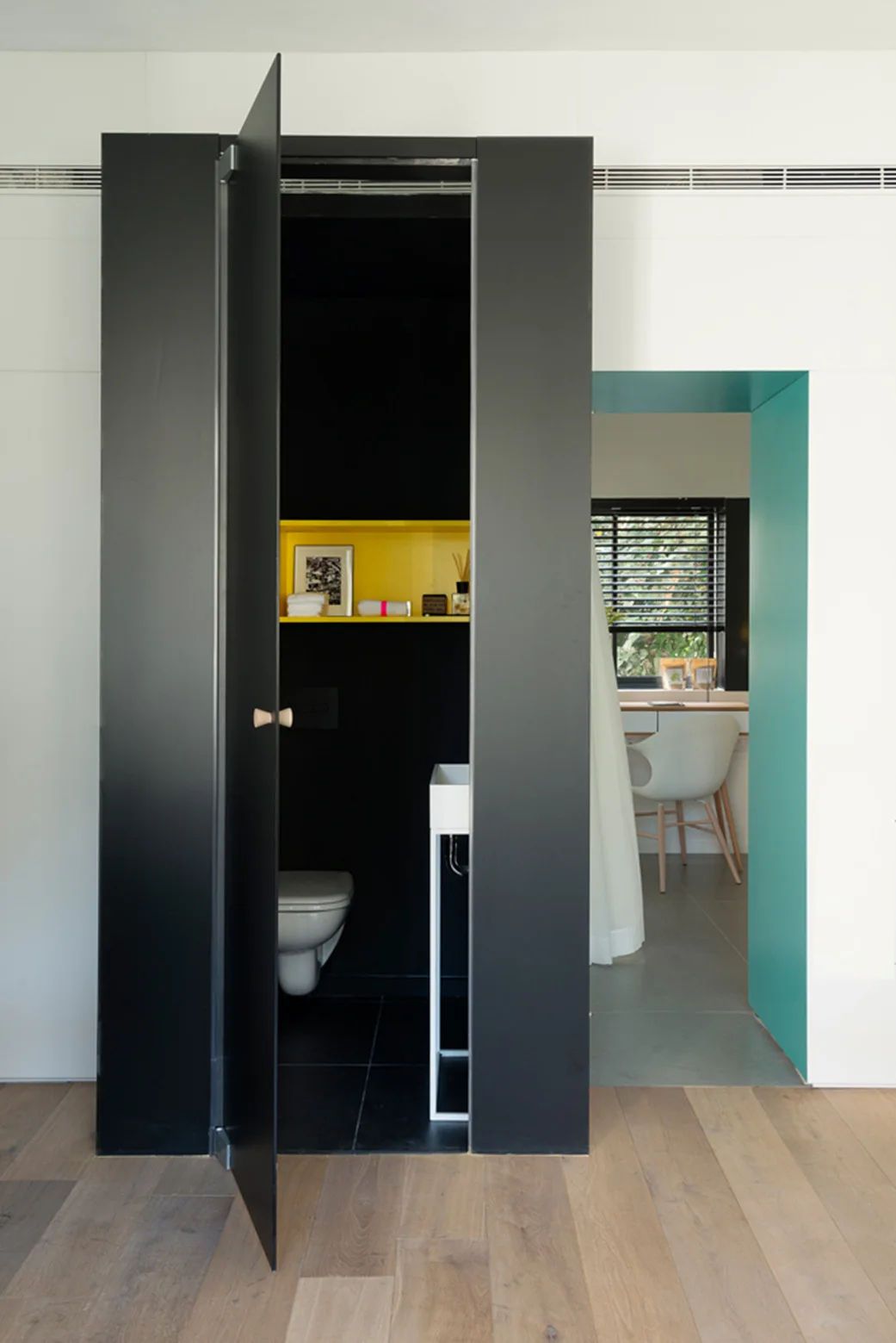 An open matte black door reveals a tiny black powder room featuring a bright yellow accent shelf next to a teal trimmed doorway.