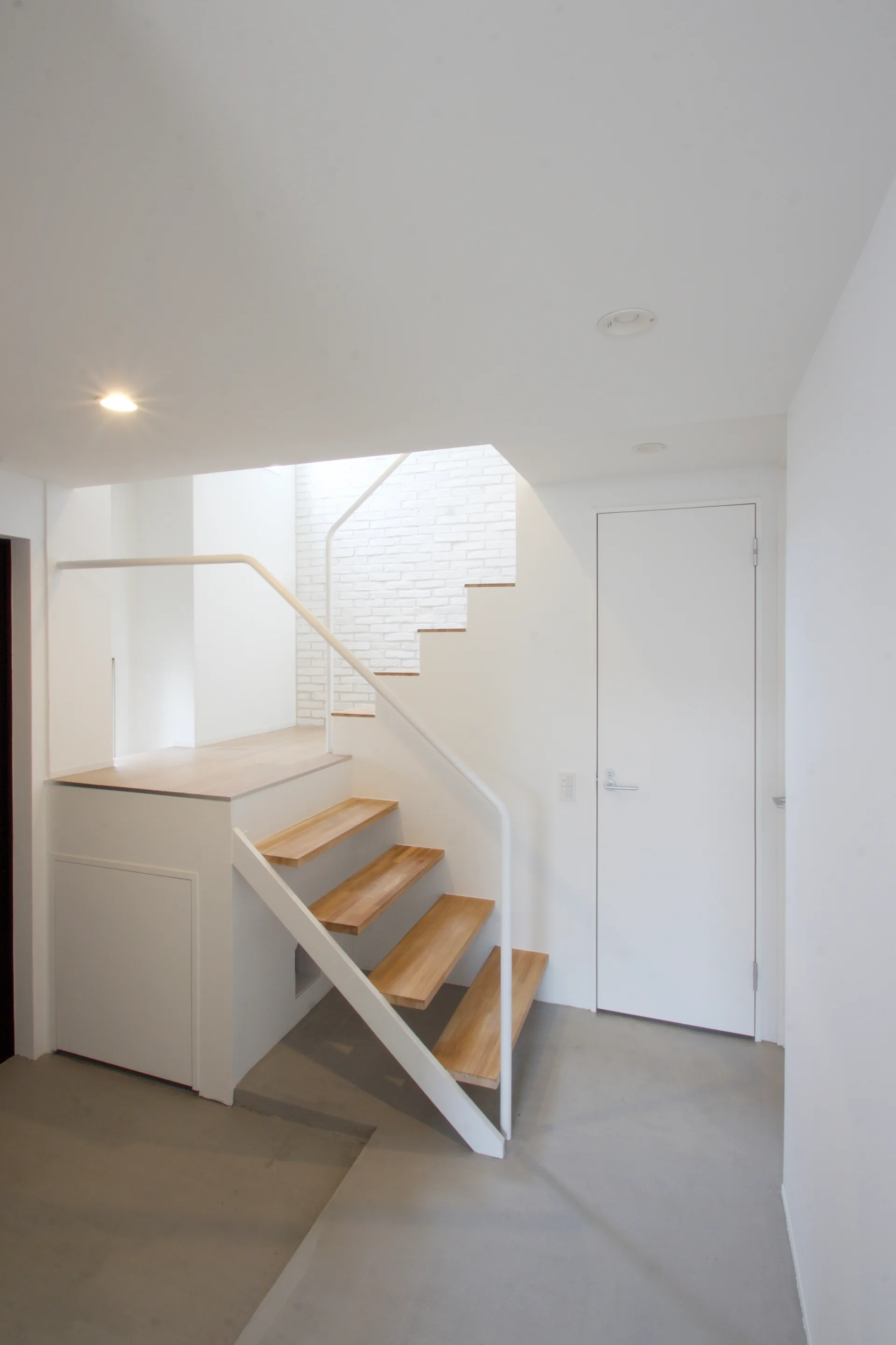 A minimalist stairwell featuring a floating wooden staircase with a white metal railing and a white painted brick wall backdrop.