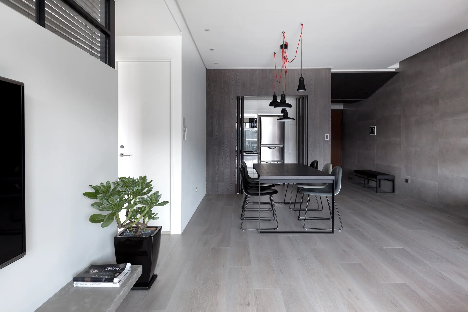 A minimalist dining room featuring a dark table and chairs set against a gray wood-paneled wall with black cluster pendant lights hung by red cords.