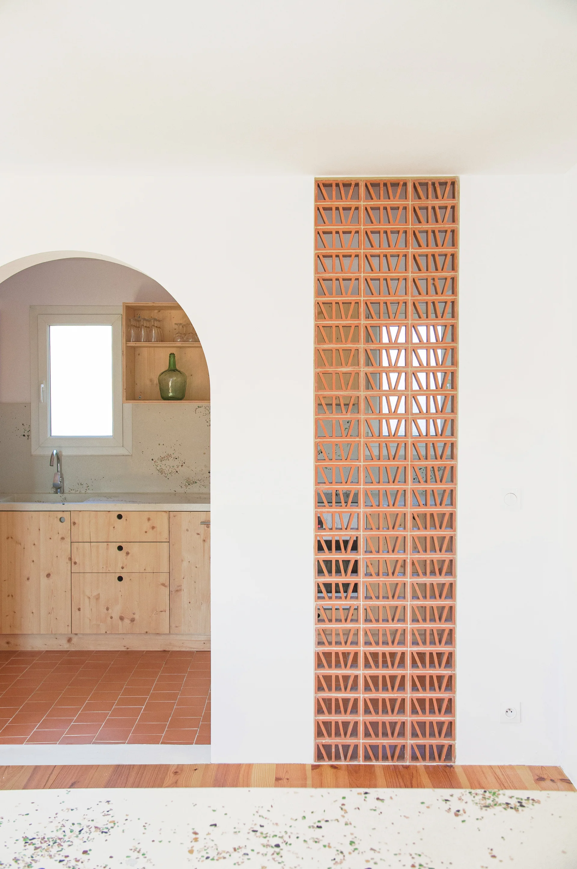 An arched doorway in a white wall reveals a kitchen with light wood cabinets and a terracotta tile floor next to a vertical breeze block screen.