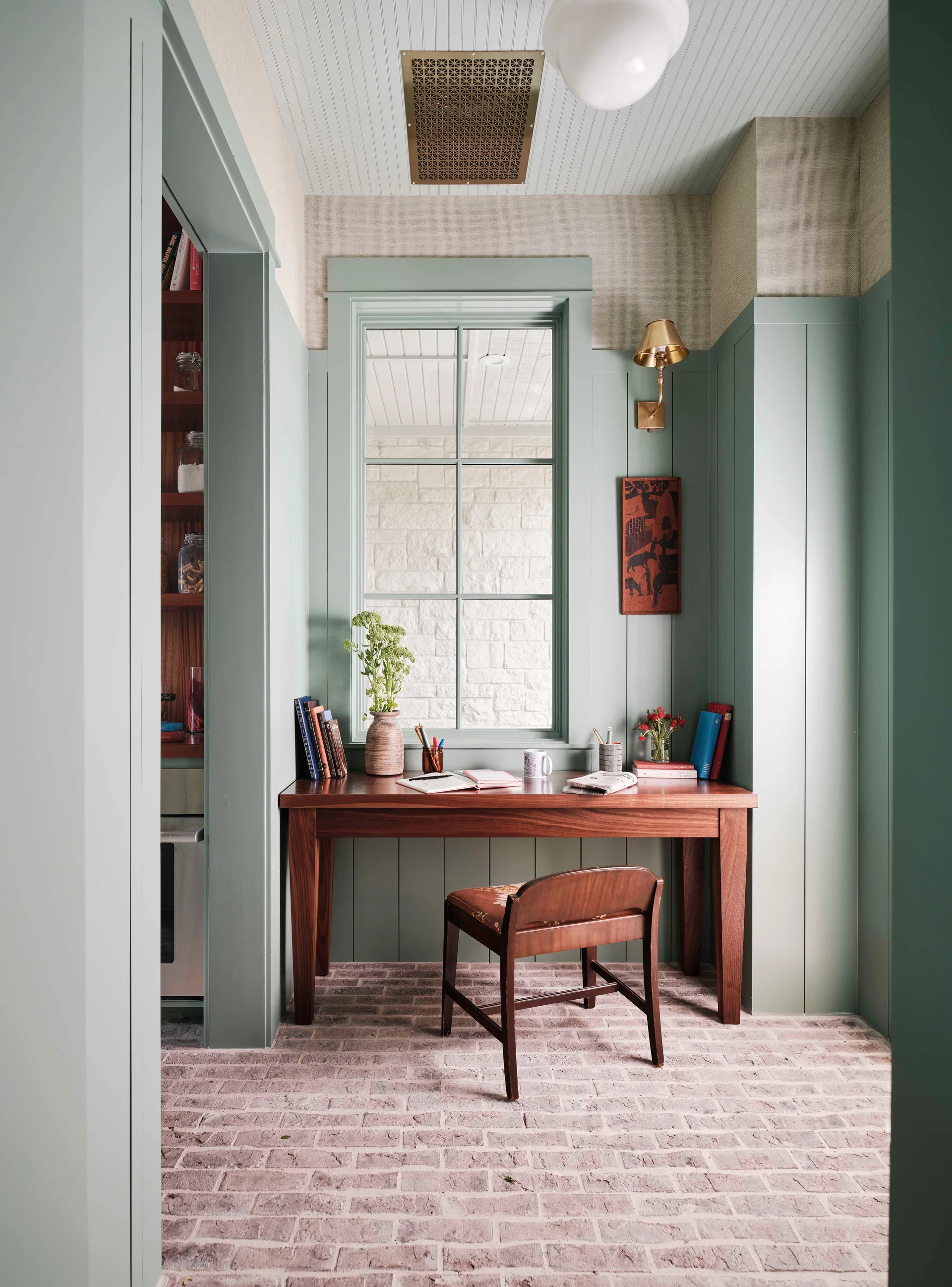 A home office alcove features a dark wood desk, sage green vertical wood paneling, and a weathered brick floor under a large window.
