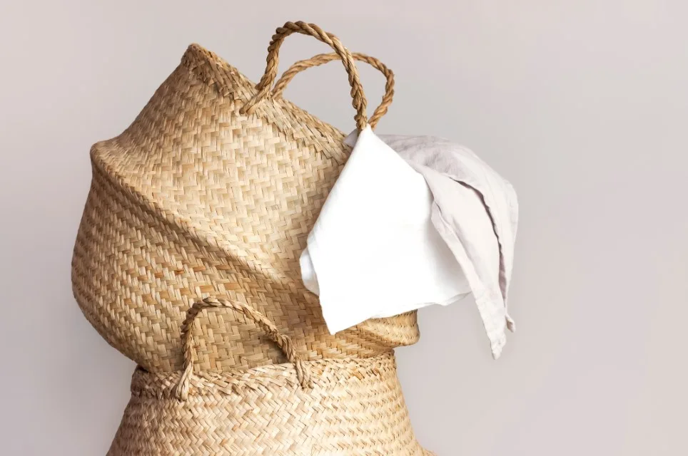 Two woven seagrass baskets are stacked against a neutral background with white and beige linen fabrics draped over the top handle.