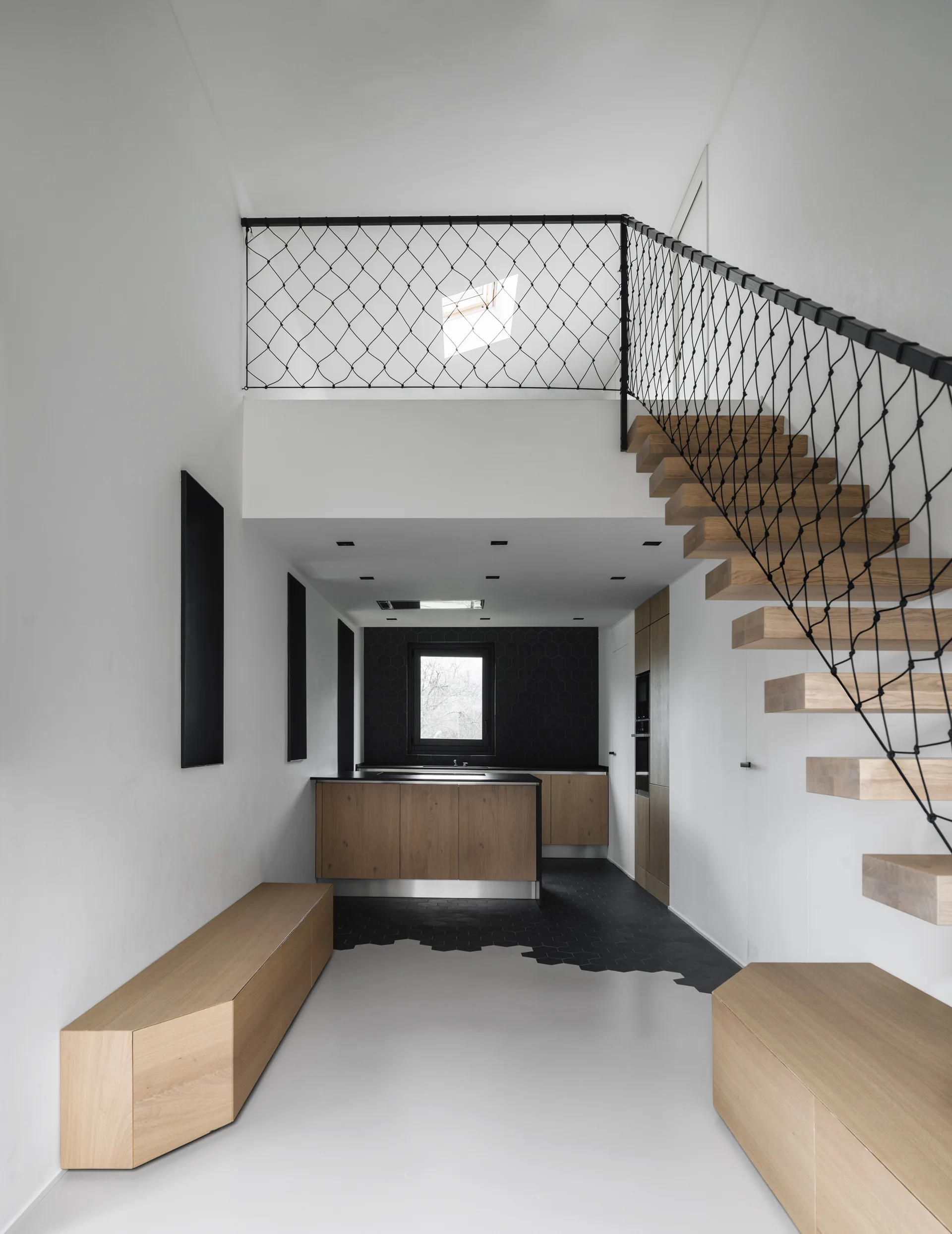 A minimalist kitchen features floating wooden stairs with black mesh netting, built-in oak cabinets, and a transition to black hexagonal floor tiles.